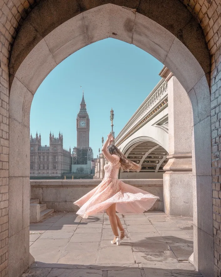 Woman in a pink twirling dress framed by a stone arch with Big Ben and Westminster Bridge in the background.