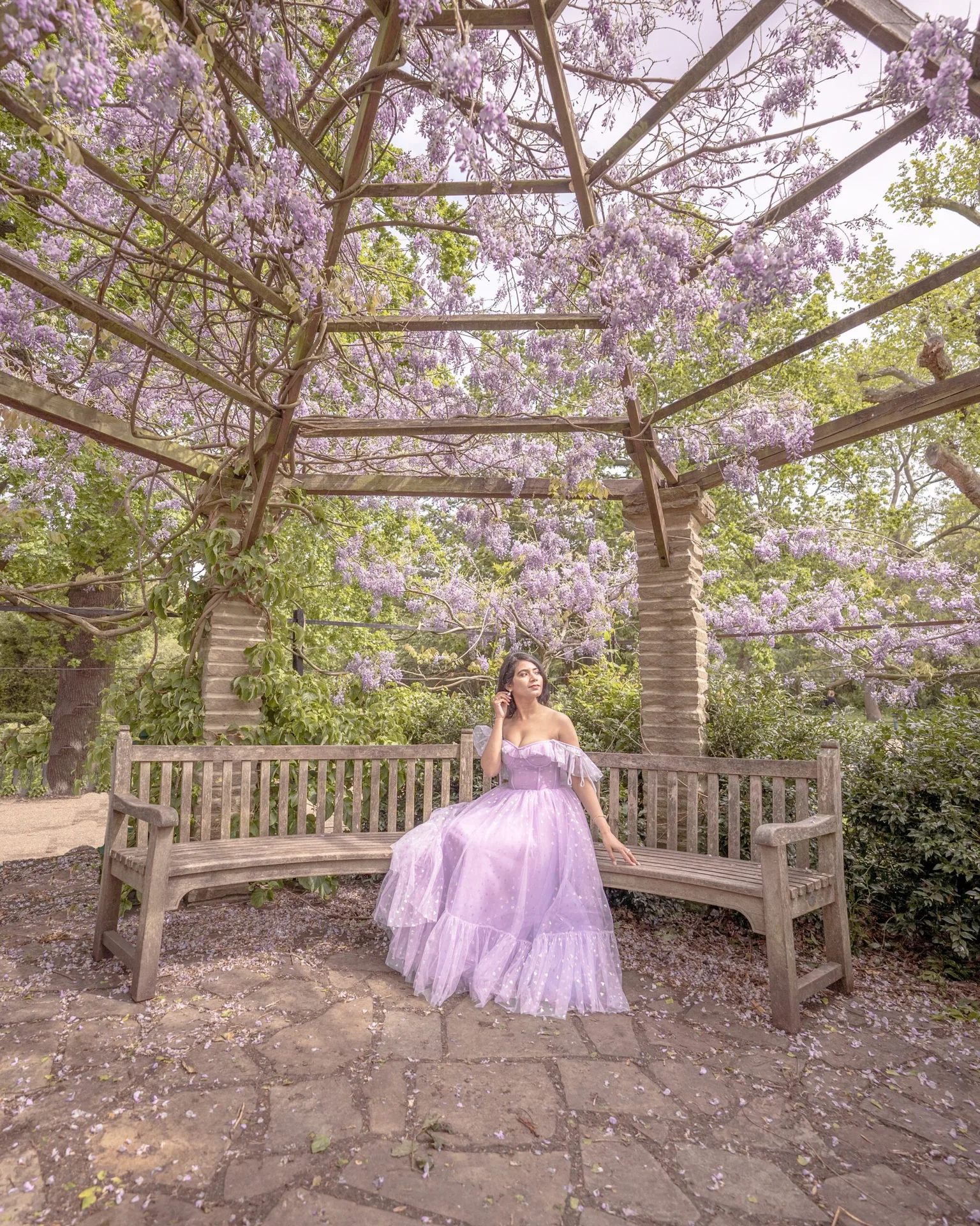 Woman in a flowing lilac gown sitting on a wooden bench beneath a pergola draped in blooming wisteria.