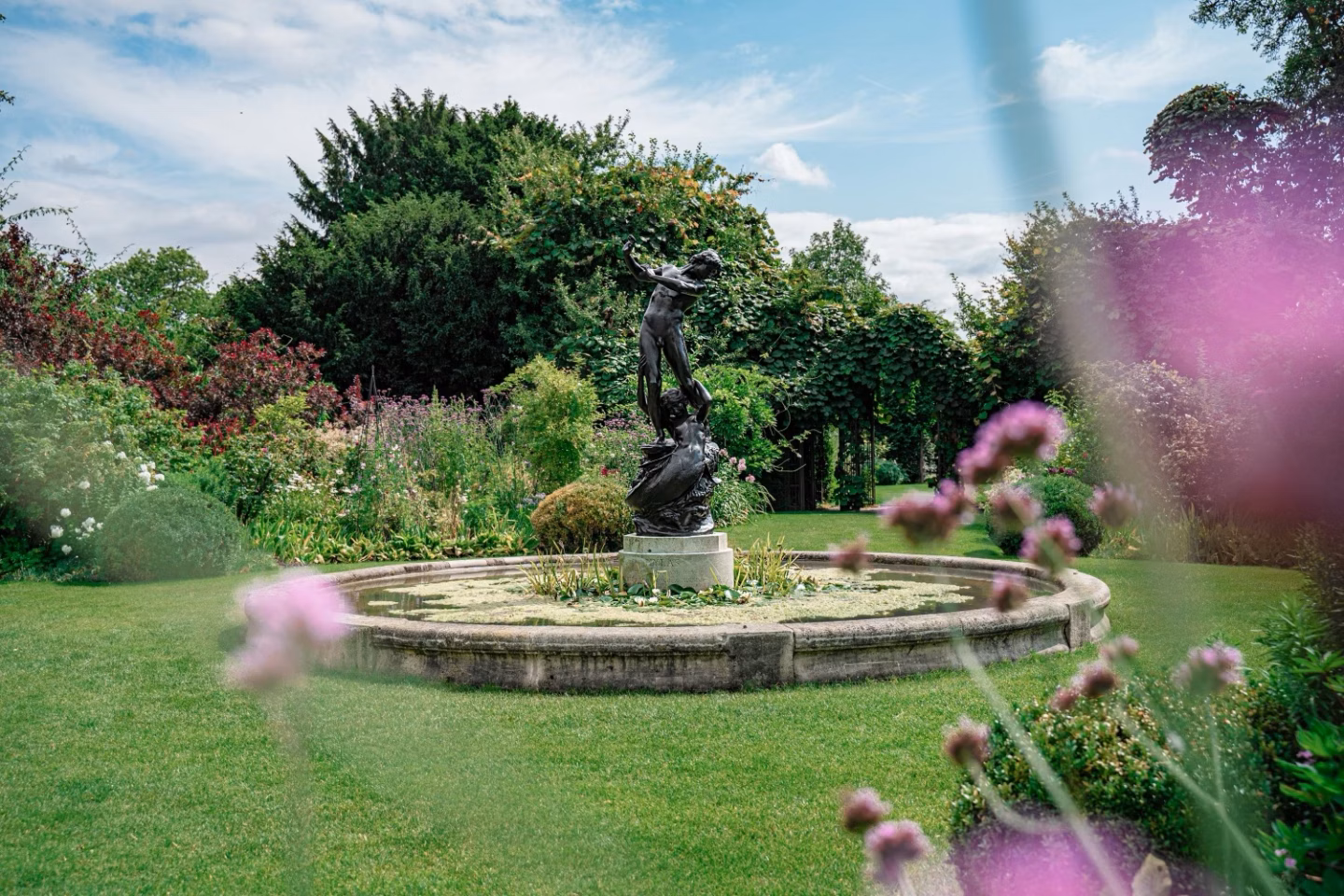 A formal garden with a bronze statue in a circular pond surrounded by lush greenery and flowers