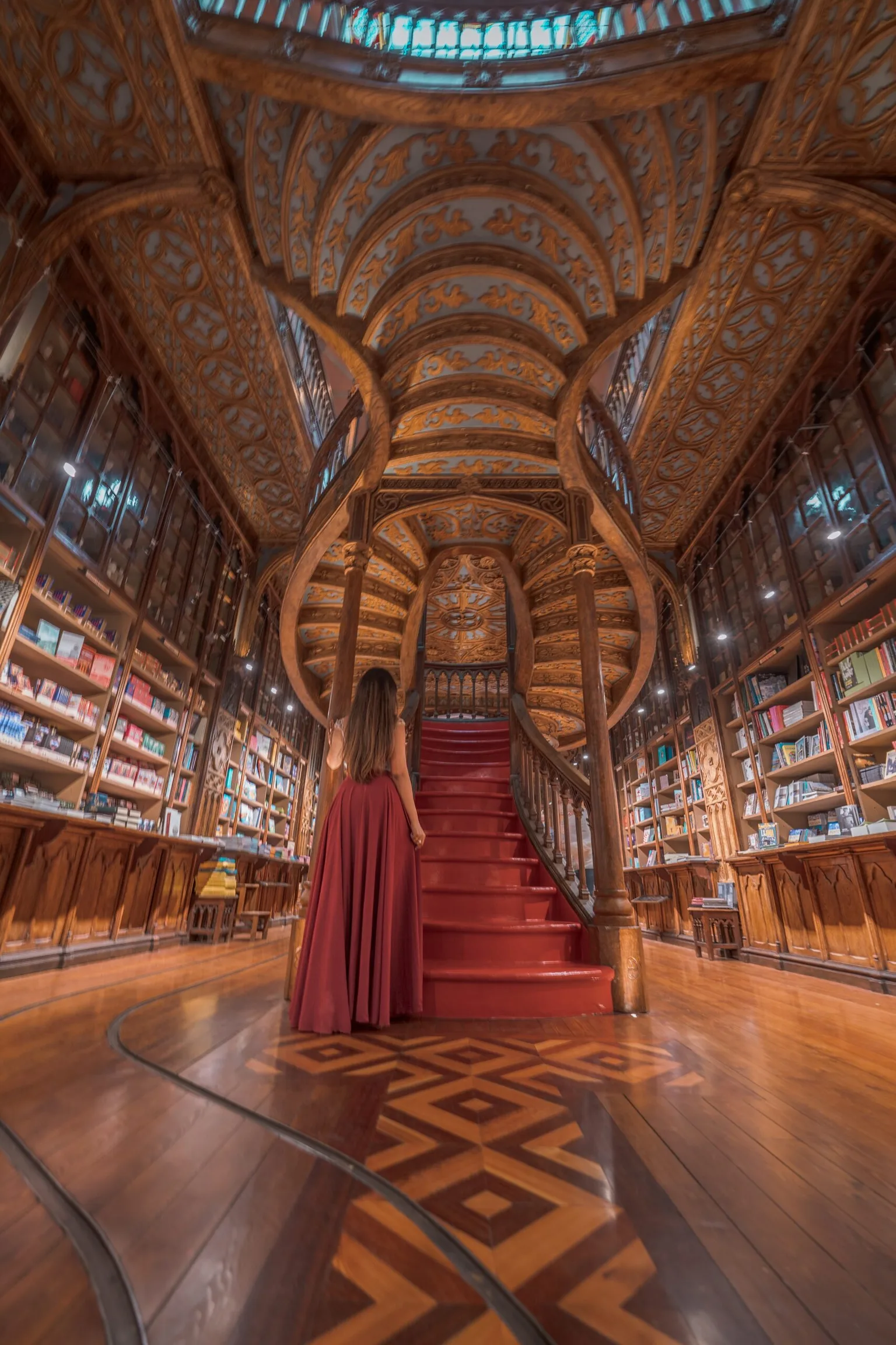 Woman in a flowing red dress ascending a carved wooden spiral staircase inside an ornate, book-lined historic library.