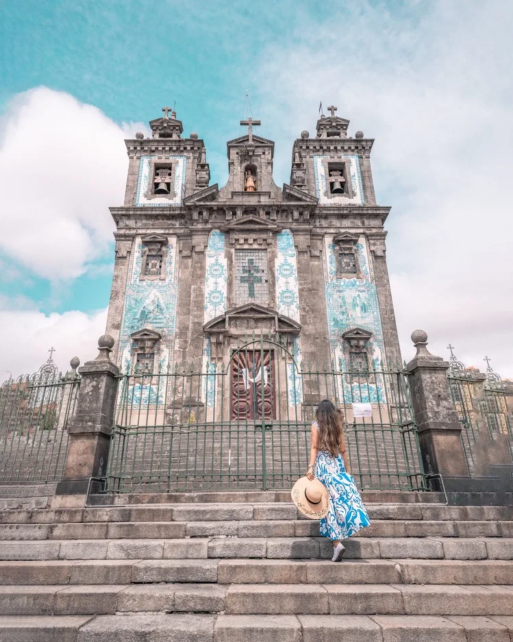 Woman in a blue patterned dress and sunhat ascending stone steps toward a historic church façade decorated with blue azulejo tiles