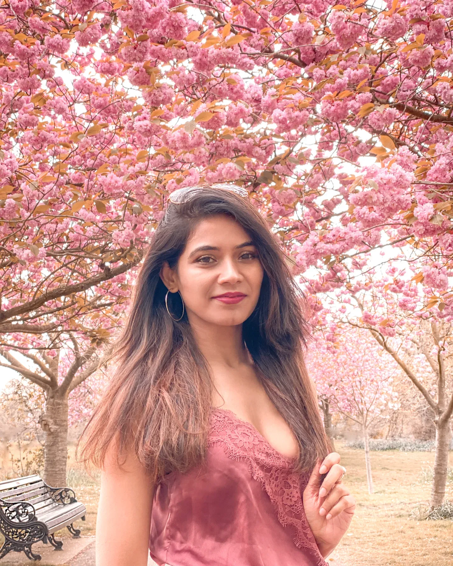 Woman smiling beneath a canopy of pink cherry blossom trees in a park, wearing a lace-trimmed rose top and hoop earrings