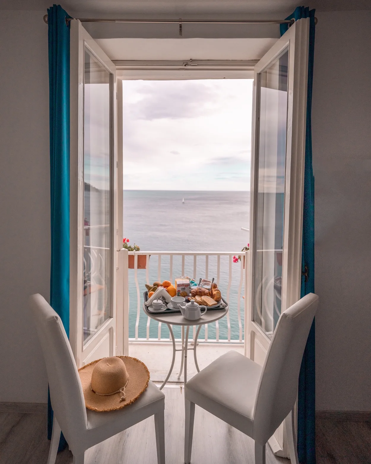 Small balcony breakfast table with croissants, fruit and tea set between two chairs overlooking a calm sea and distant sailboat.