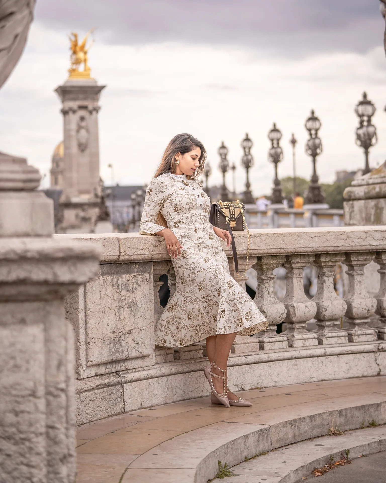 Woman in a floral midi dress leaning against an ornate stone balustrade on a Parisian bridge with lamp posts and gilded statue in the background.