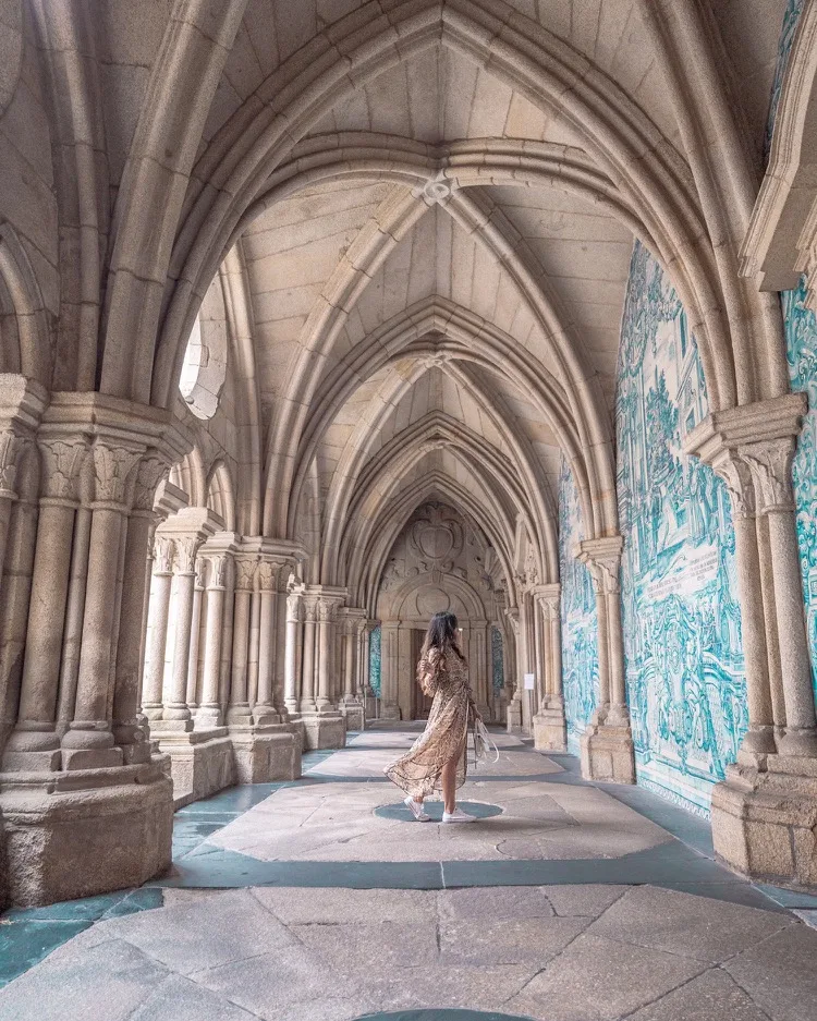 Woman in a flowing dress strolling through a vaulted stone cloister lined with blue azulejo tile murals.