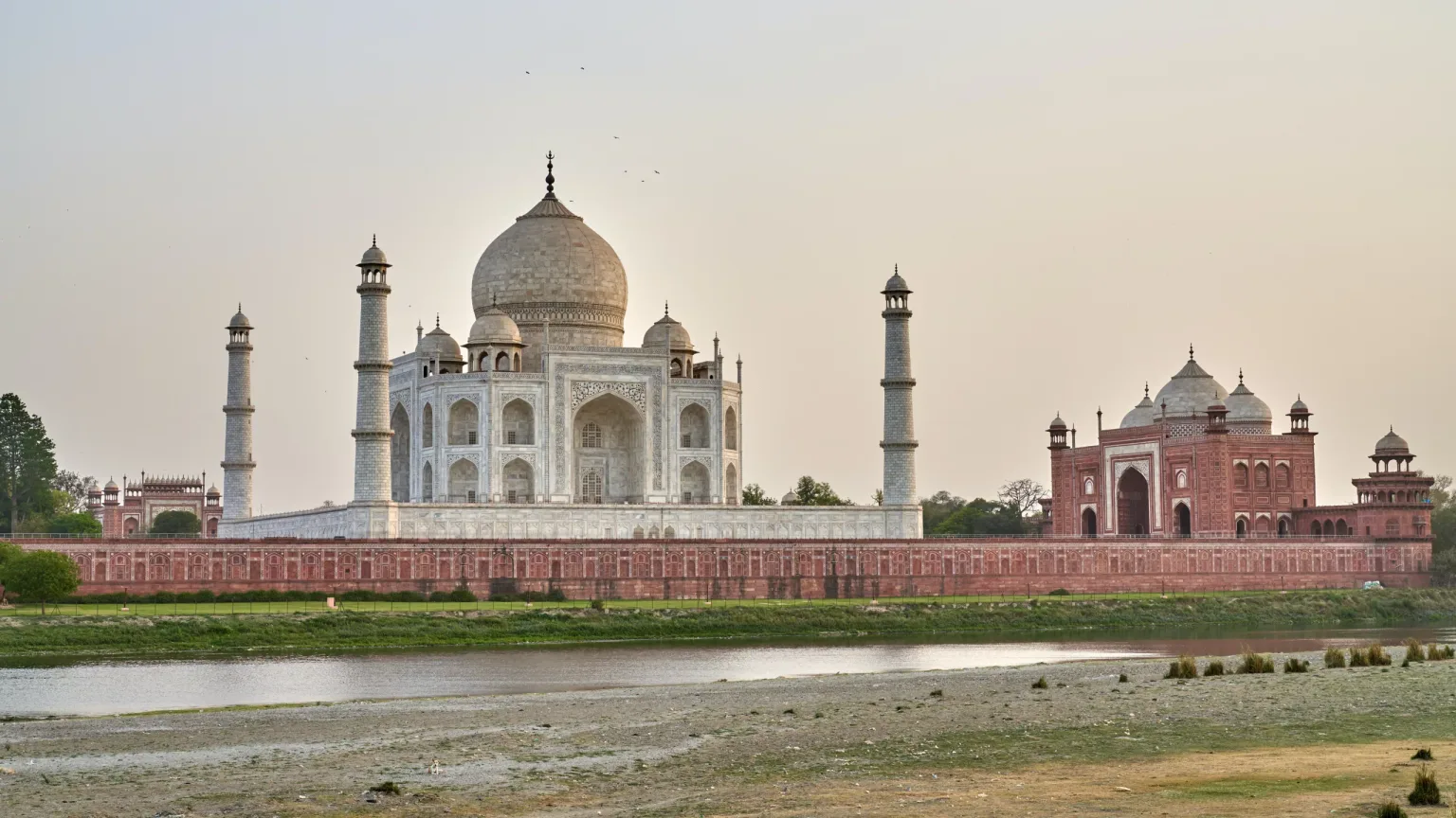 The white marble Taj Mahal with its central dome and four minarets reflected beside the Yamuna River at sunset.
