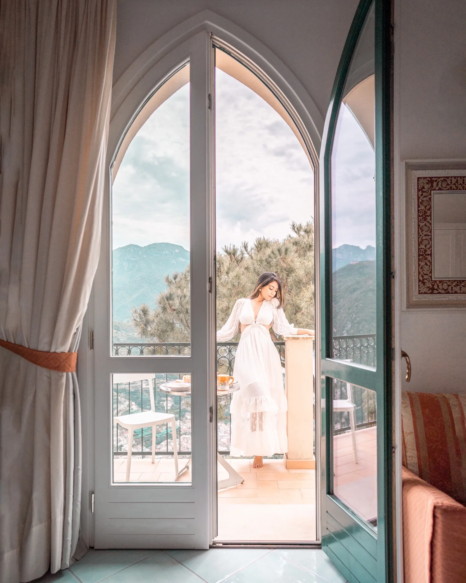Woman in a flowing white dress standing on a sunlit balcony overlooking mountains and a town, seen through arched glass doors.