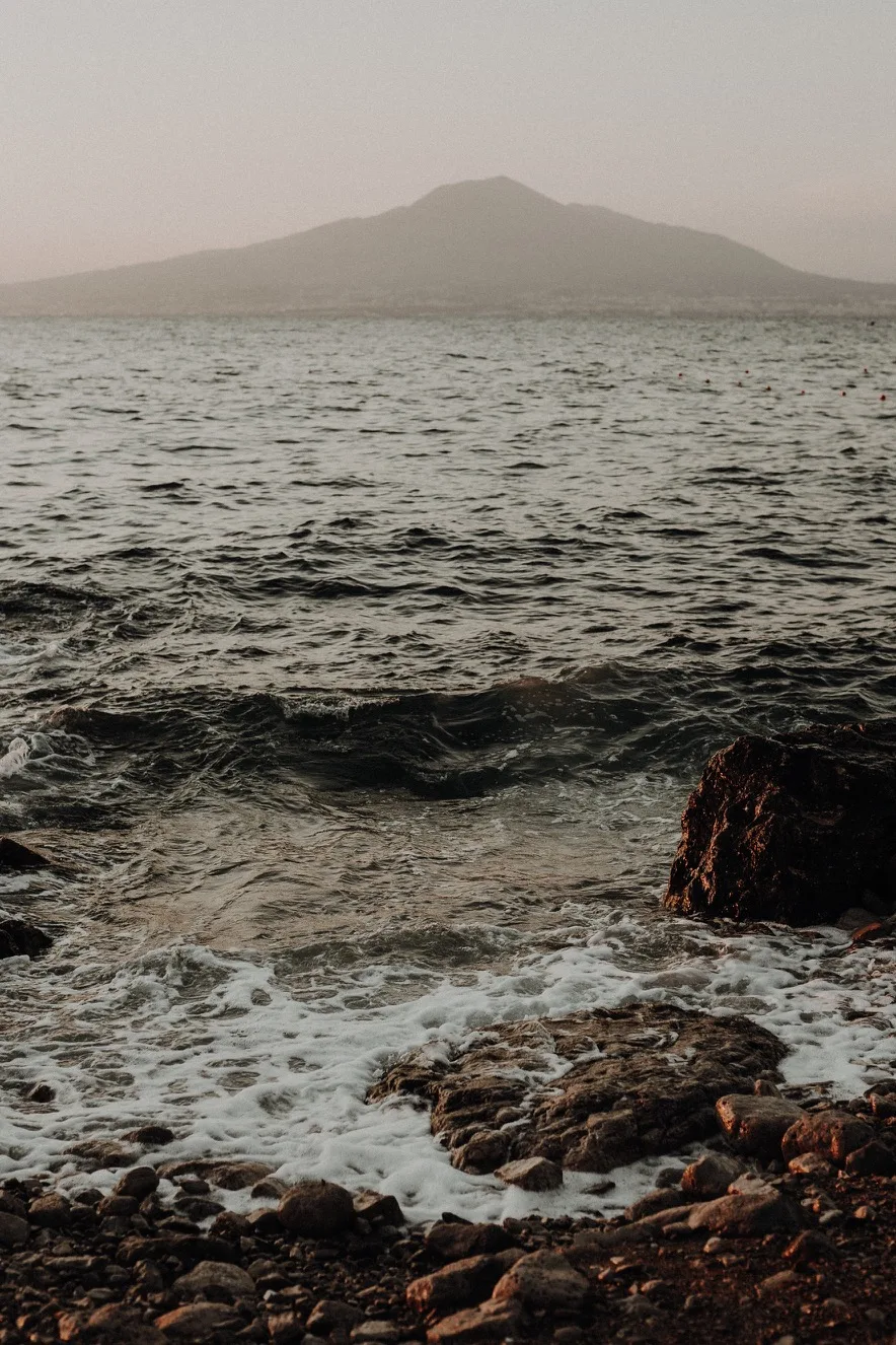 Rocky shoreline with foaming waves in the foreground and a hazy volcanic island silhouette on the horizon at dusk.