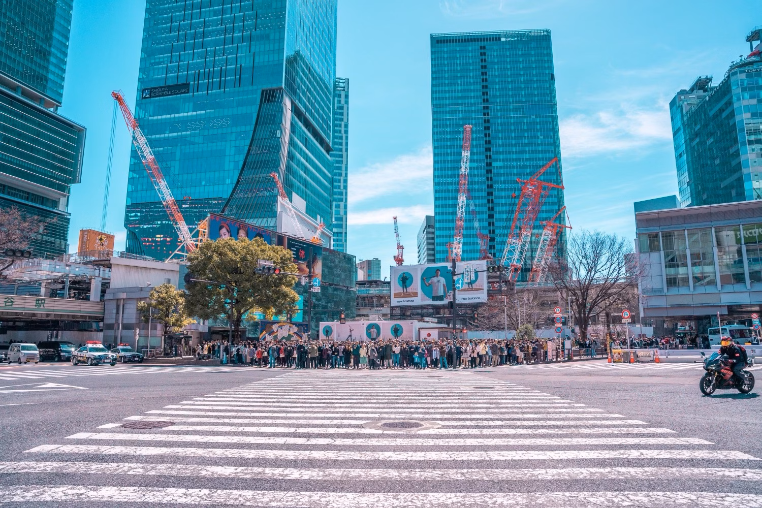 Busy city intersection with a wide zebra crossing, crowds, and modern glass towers under construction