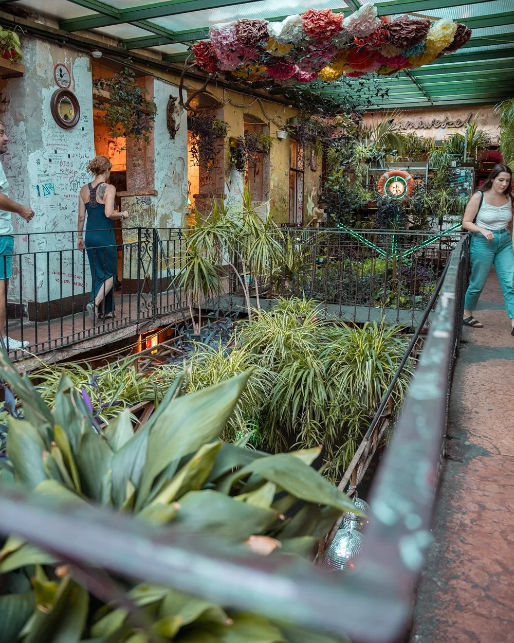 Indoor courtyard cafe with hanging paper flowers, lush tropical plants and people walking along a wrought-iron balcony.