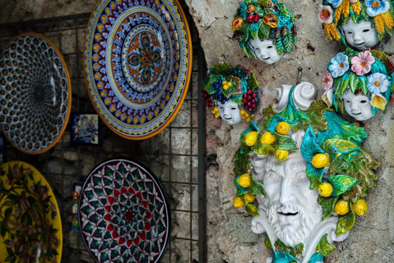 Decorative ceramic plates and colourful Sicilian-style wall masks, including a large lemon-adorned bearded face, displayed on a stone wall.