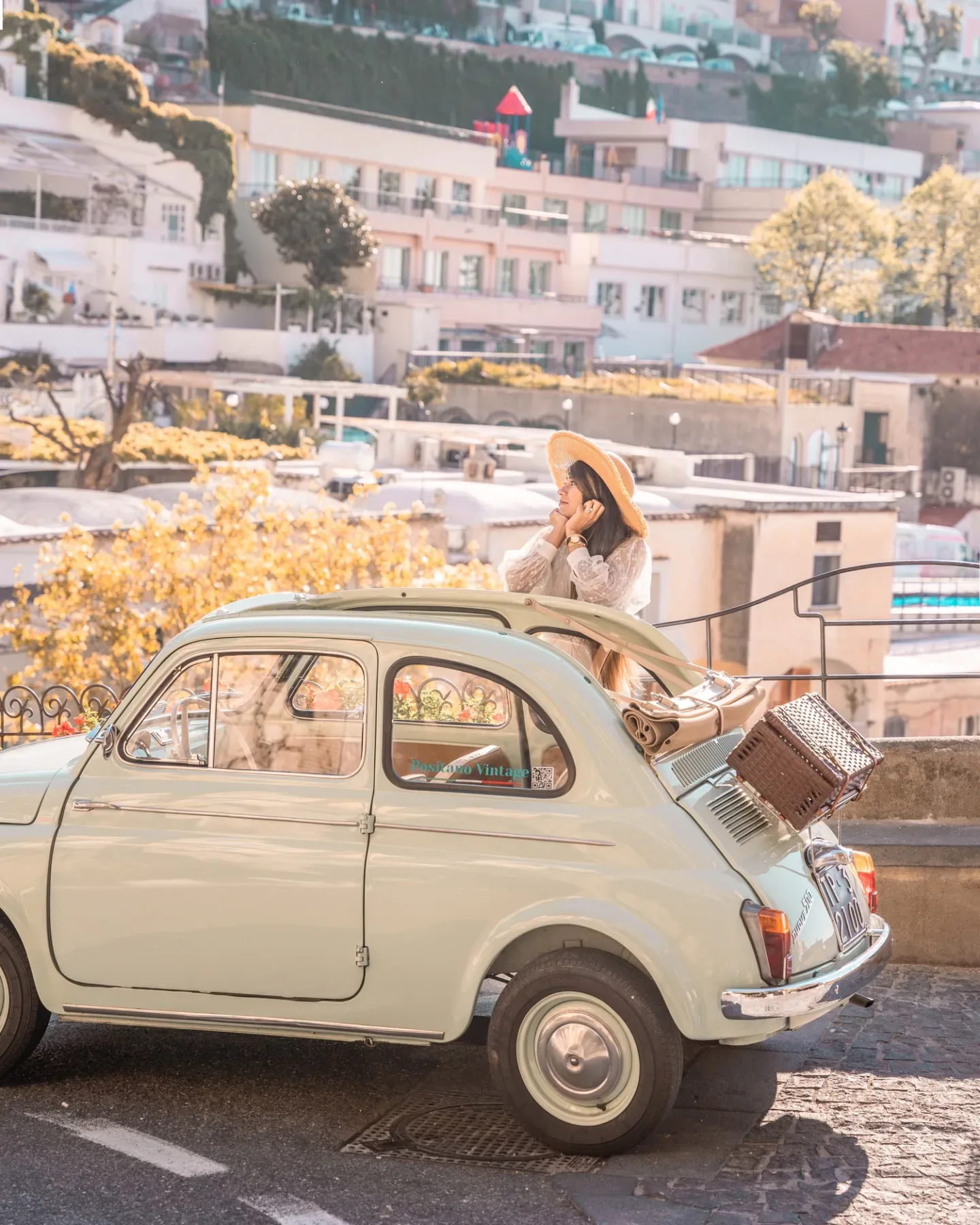Best Instagram spots Amalfi Coast - Woman in a sun hat leaning on a pale vintage Fiat with a picnic basket on the trunk, overlooking a sunlit Mediterranean hillside town.