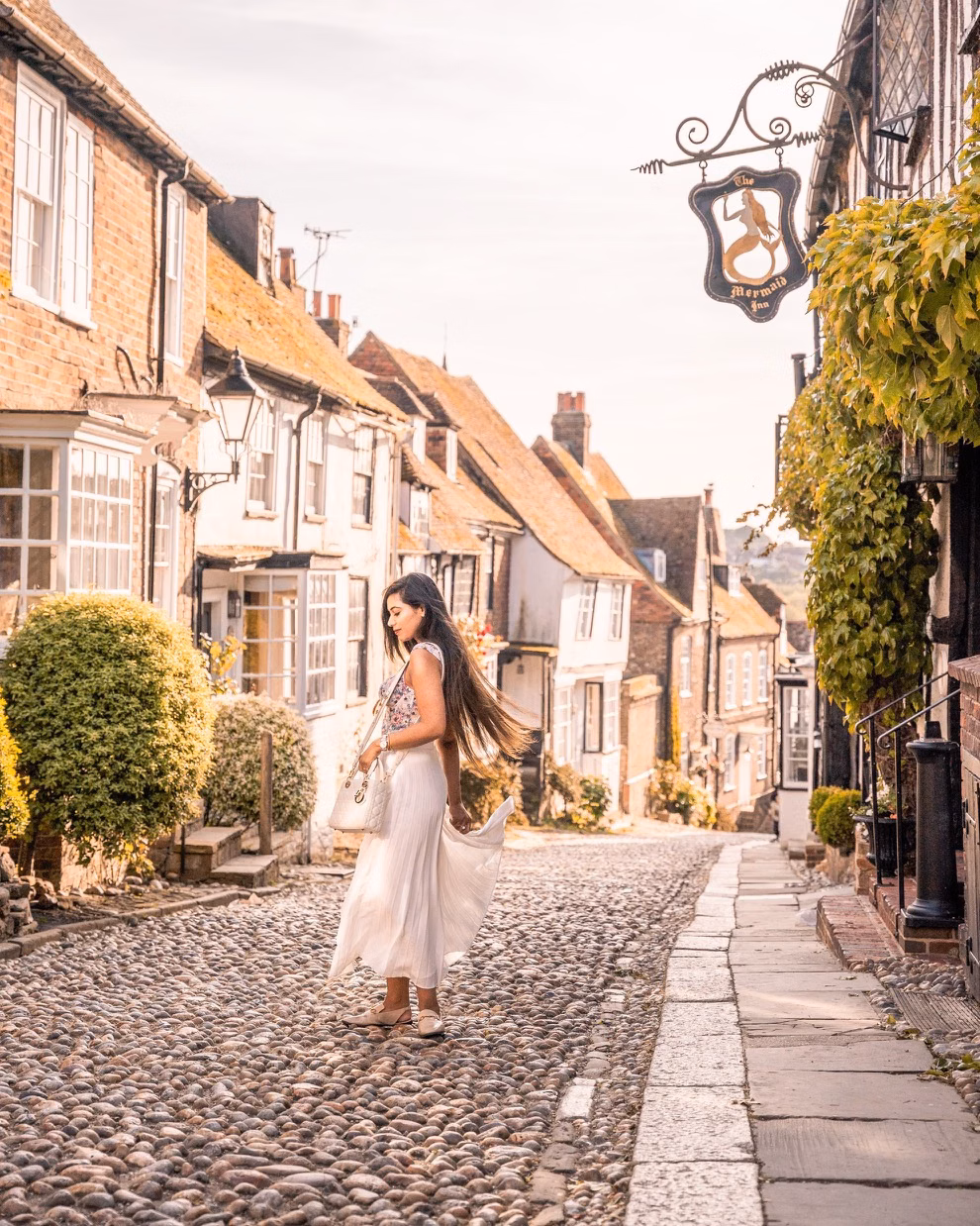 A woman in a flowing white dress walks along a cobbled street lined with historic houses