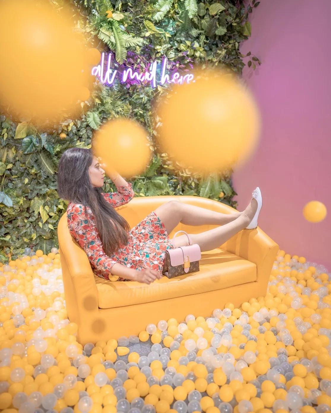 Woman in a floral dress reclining on a yellow sofa surrounded by yellow and clear ball-pit balls with a neon sign and leafy wall behind.