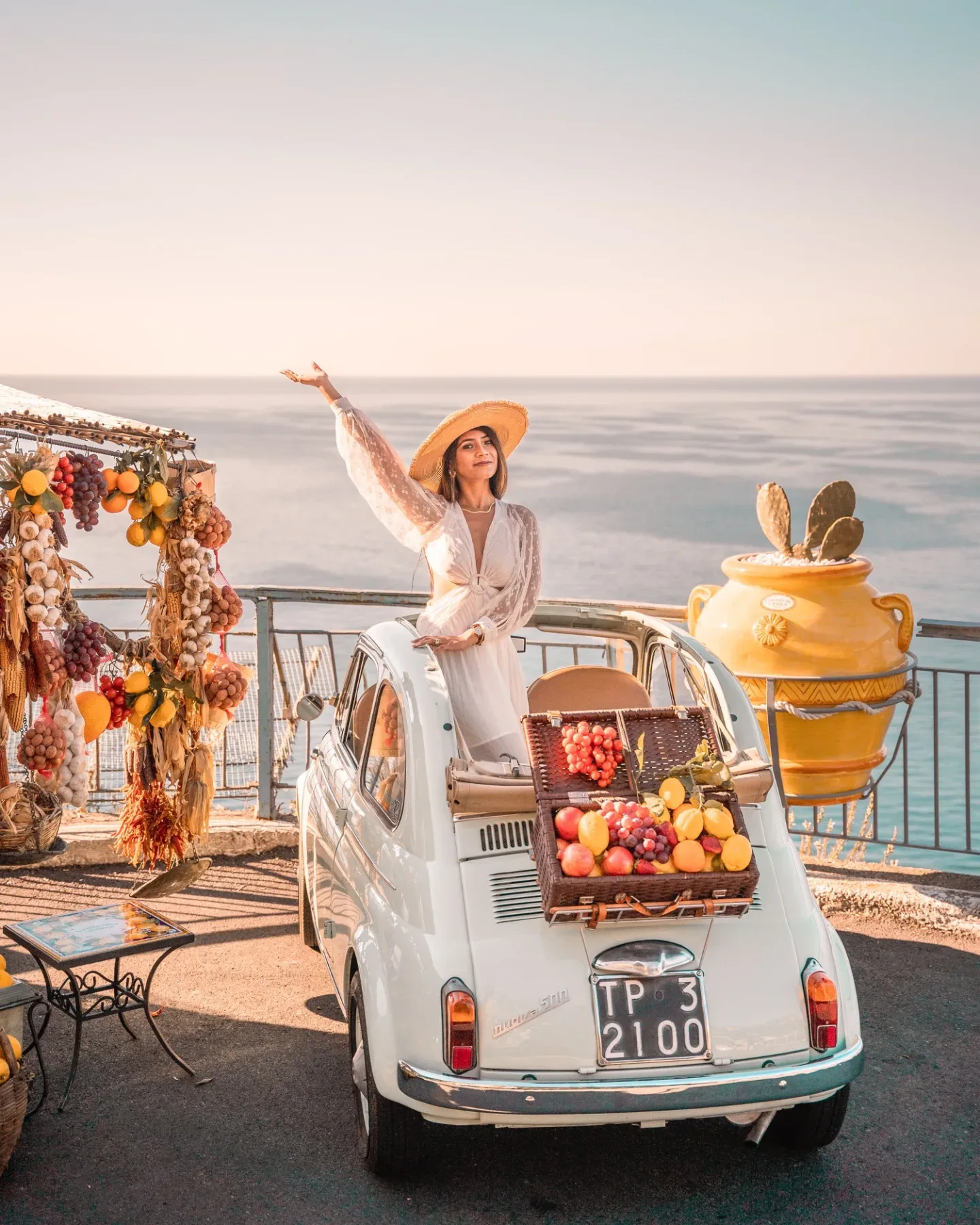 Woman in a sun hat standing through the roof of a vintage white Fiat 500 parked on a coastal overlook with a picnic basket of fruit and hanging produce displays nearby