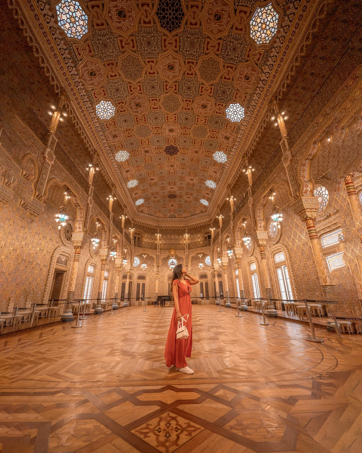 Woman in a flowing coral dress standing on an ornate parquet floor beneath an intricately patterned gilded ceiling in a grand Moorish-style palace hall