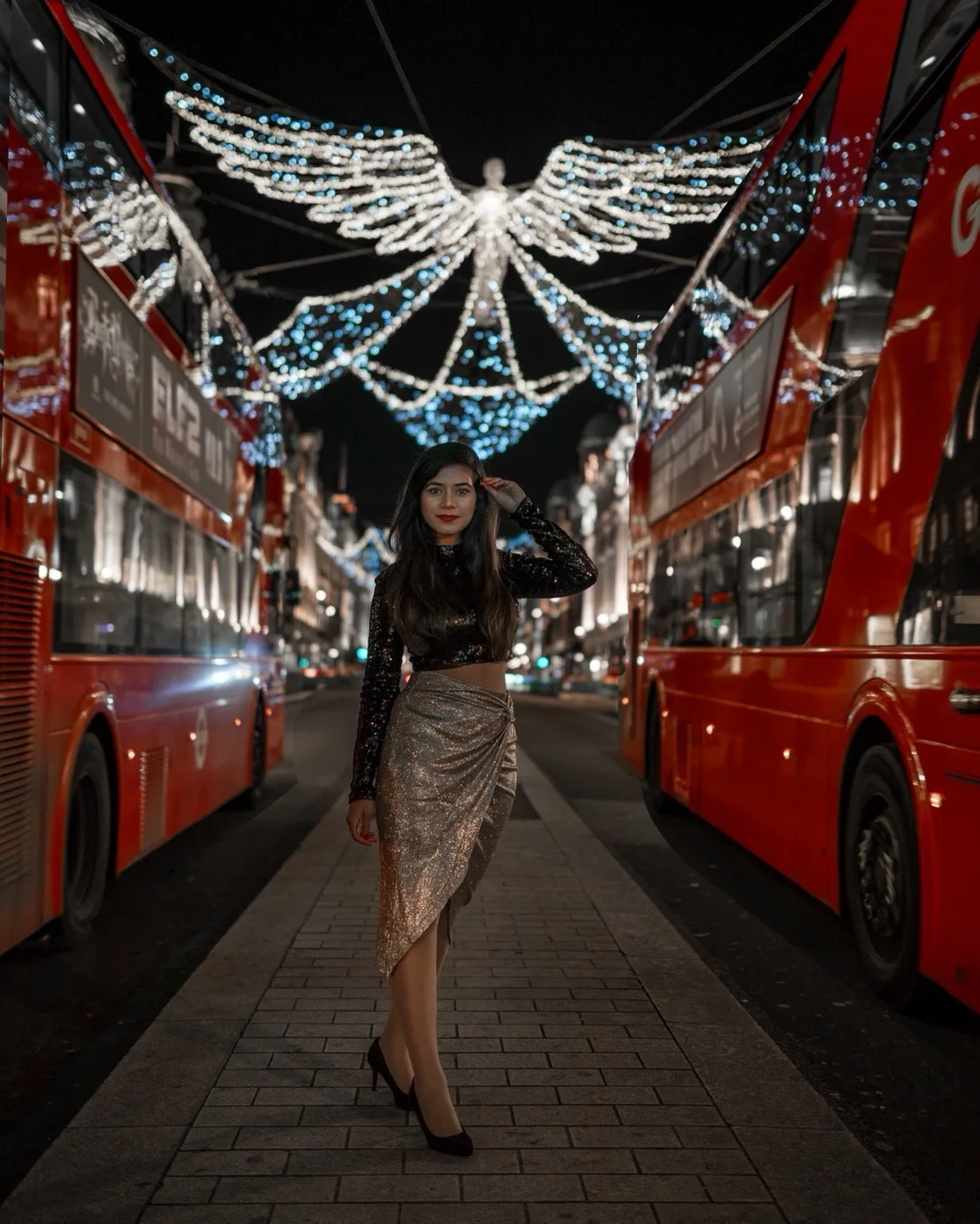 Woman in a sequined top and metallic skirt posing between two red double-decker buses under illuminated angel street lights at night.