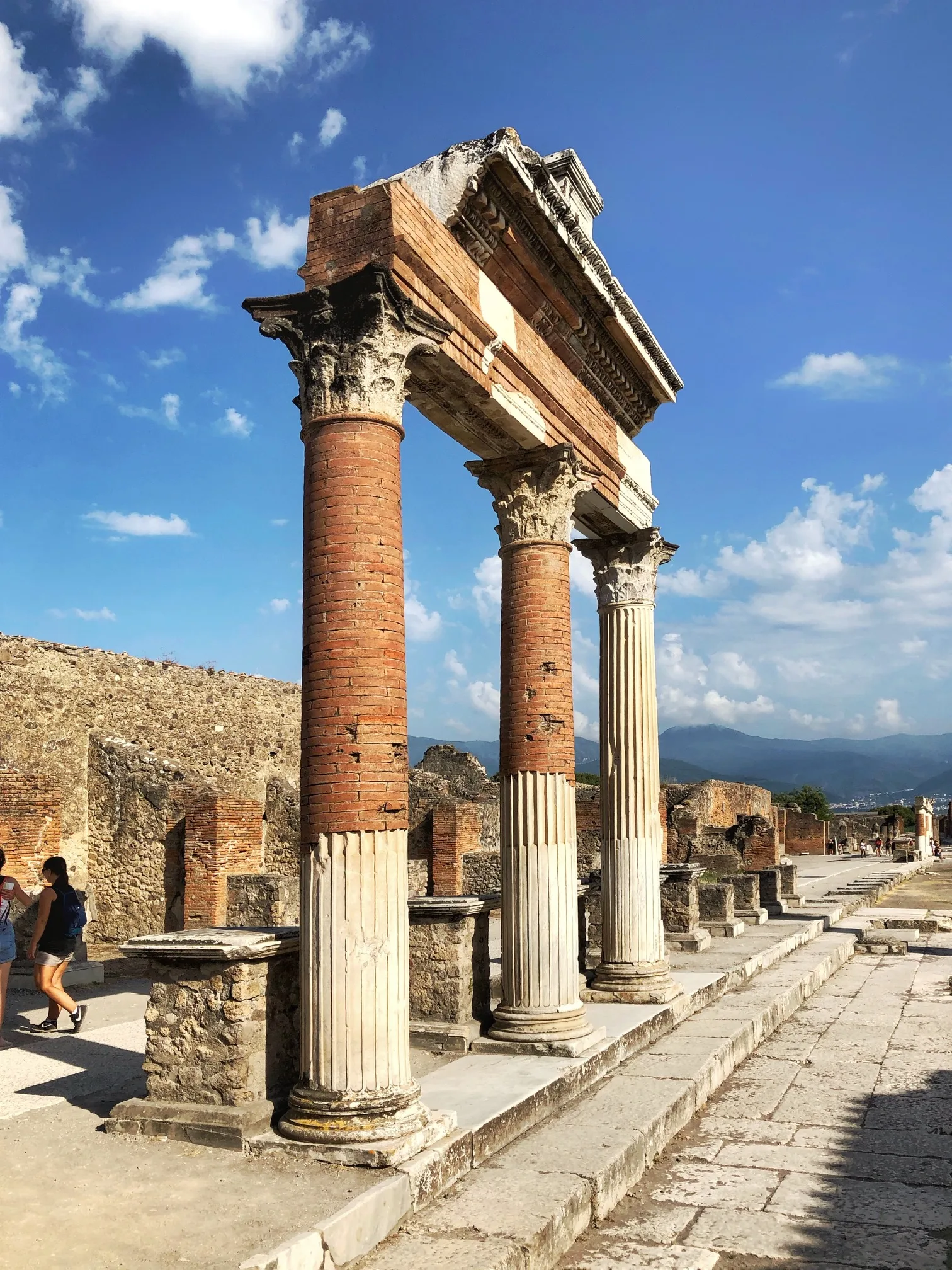 Ancient Roman brick and marble columns and a ruined portico standing along a sunlit paved street at Pompeii with mountains and blue sky in the background