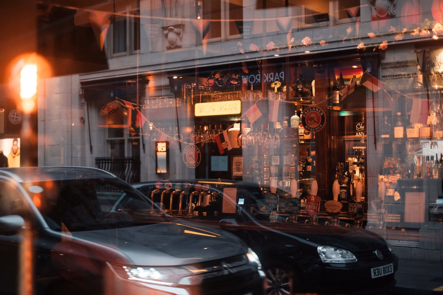 A warmly lit pub front at dusk, seen through a reflective window with parked cars in the foreground