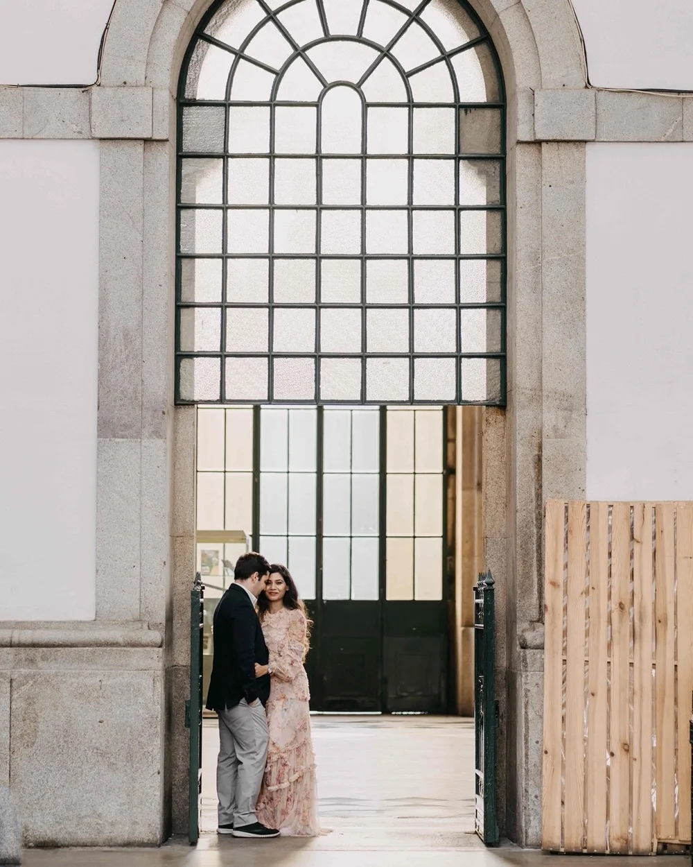 Couple standing intimately beneath a tall arched window in a sunlit stone doorway, the woman in a flowing floral dress and the man in a dark jacket.