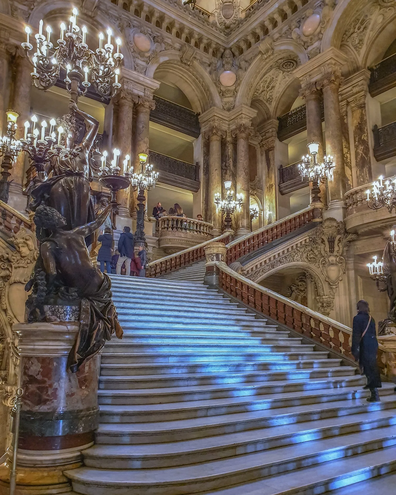 Grand marble staircase with ornate bronze candelabras, carved balustrades and glowing chandeliers inside a lavish historic opera house.