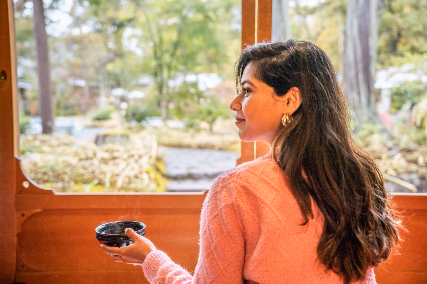 Woman in a pink sweater holding a bowl and gazing out a train window at a blurred green landscape