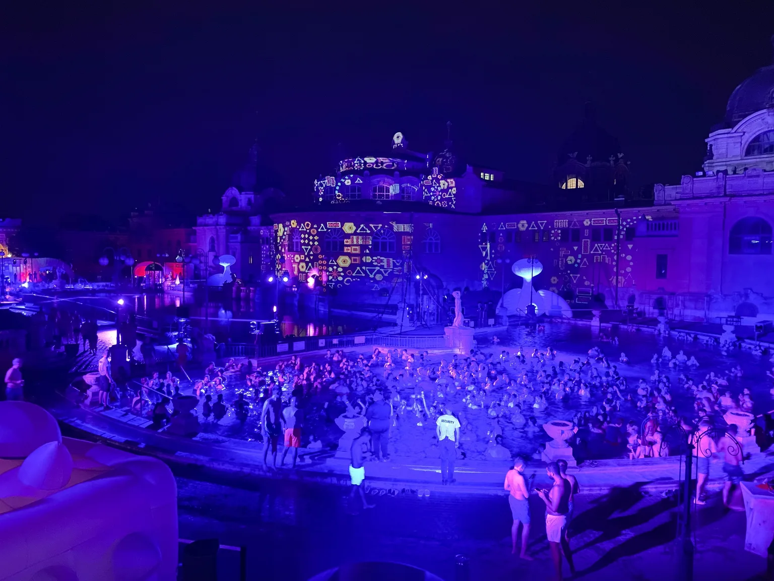 Crowd of people enjoying a night-time light show and pool party at a historic thermal bath complex lit in vivid purple and projected patterns.