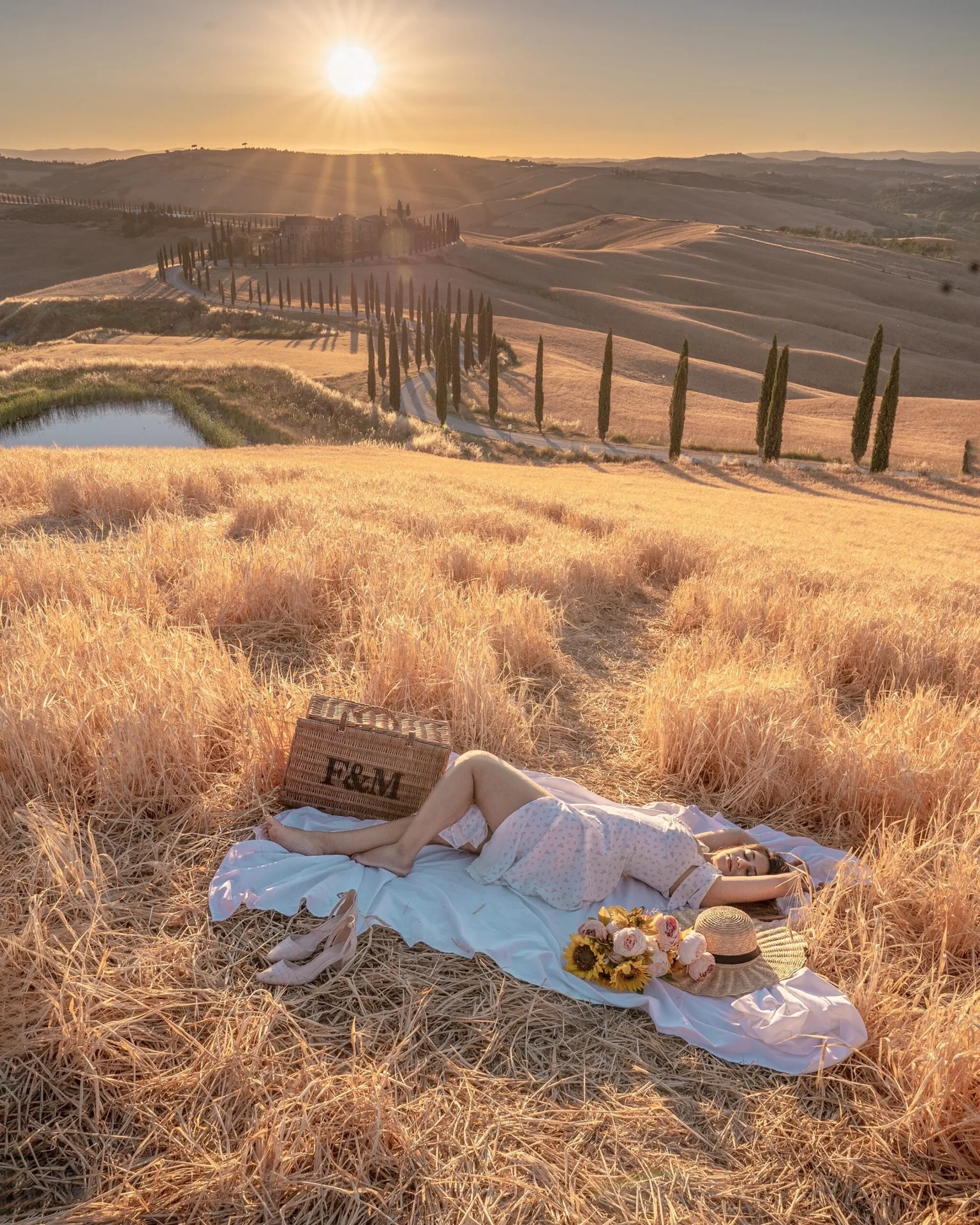 Woman lying on a picnic blanket with a wicker basket, hat and flowers in golden rolling hills at sunset, cypress-lined road in the distance