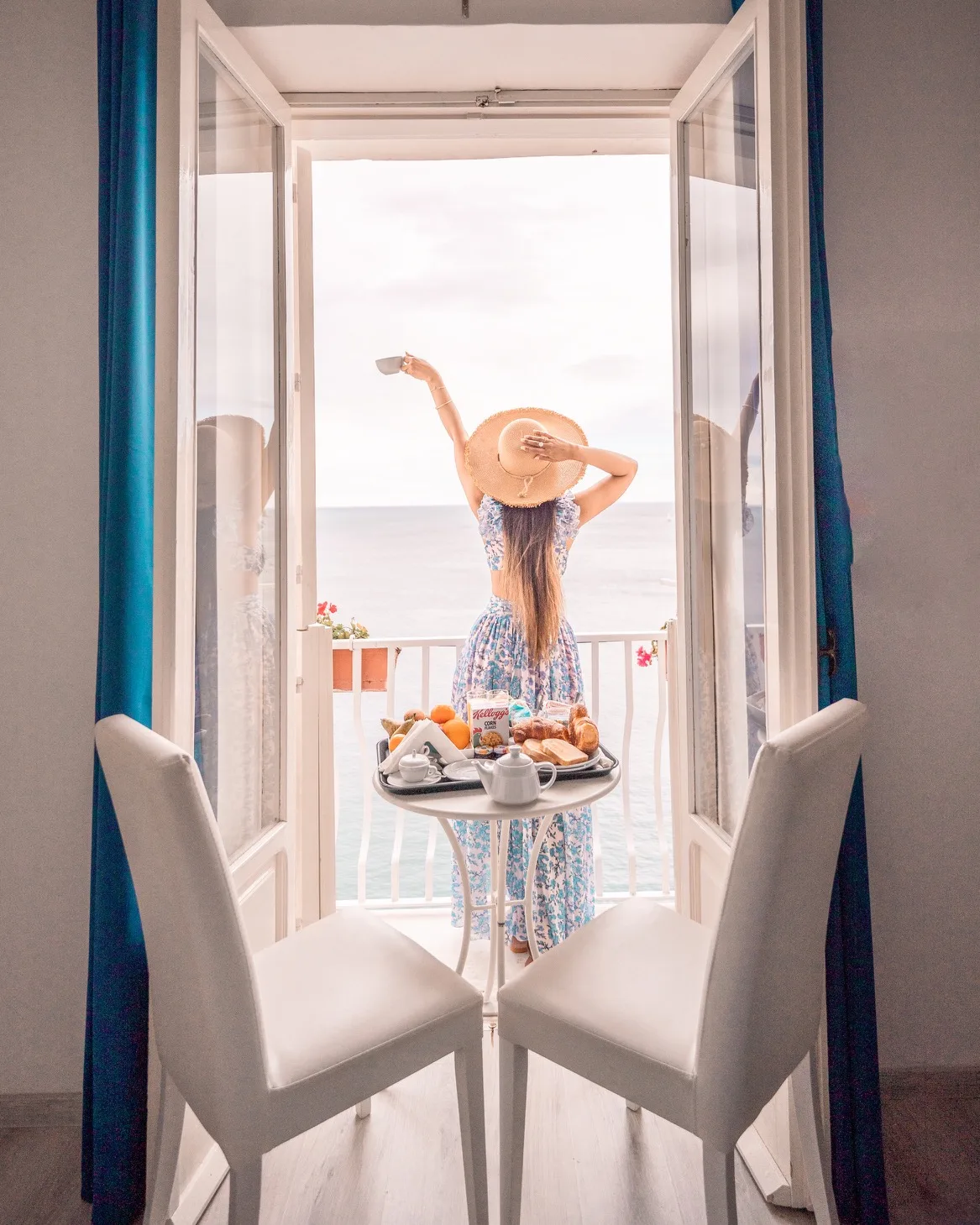 Amalfi Woman in a sunhat raises a cup on a balcony overlooking the sea with a table set for breakfast.