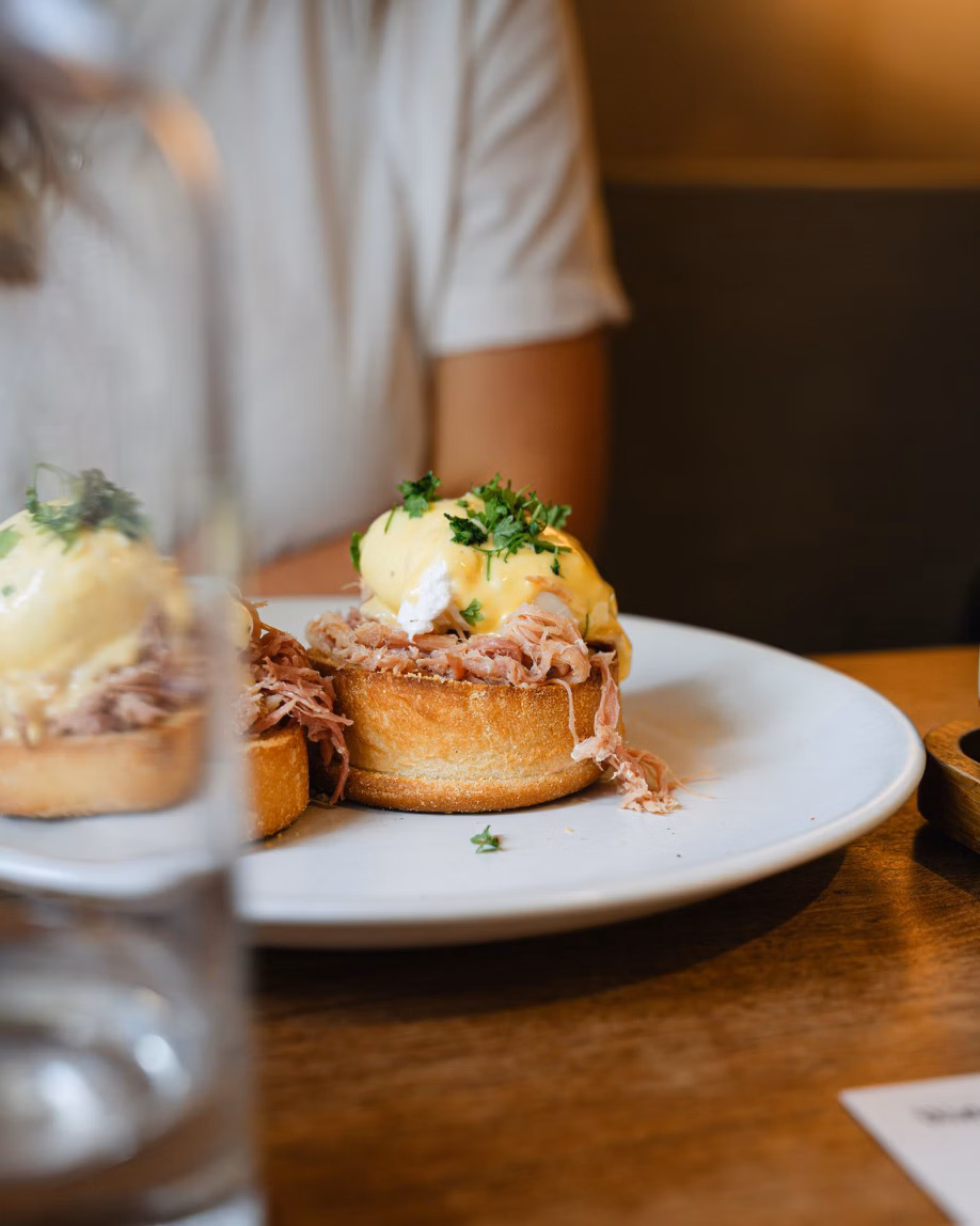 Eggs Benedict on toasted muffins with pulled meat, served on a white plate in a cosy restaurant