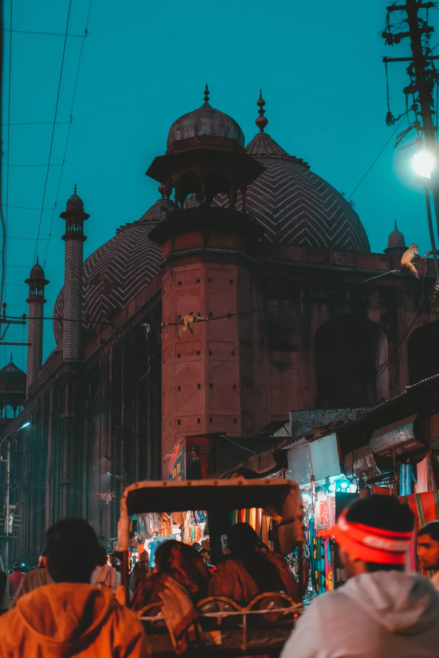 Crowded evening market street with rickshaws and shoppers beneath the lit domes and minarets of an ornate mosque.