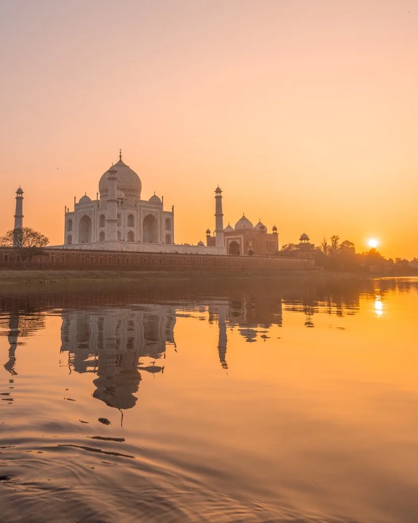 Taj Mahal silhouetted against a golden sunrise with its reflection mirrored in the calm Yamuna River.