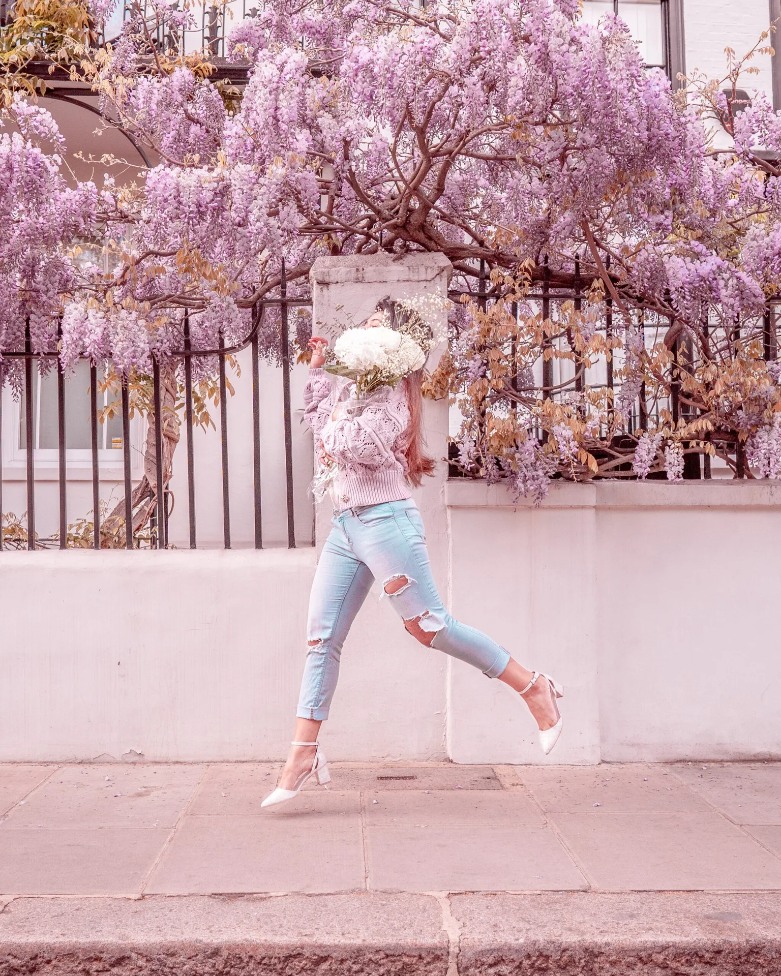 Woman mid-leap along a sidewalk holding a large bouquet beneath cascading lilac wisteria on a pastel city street.