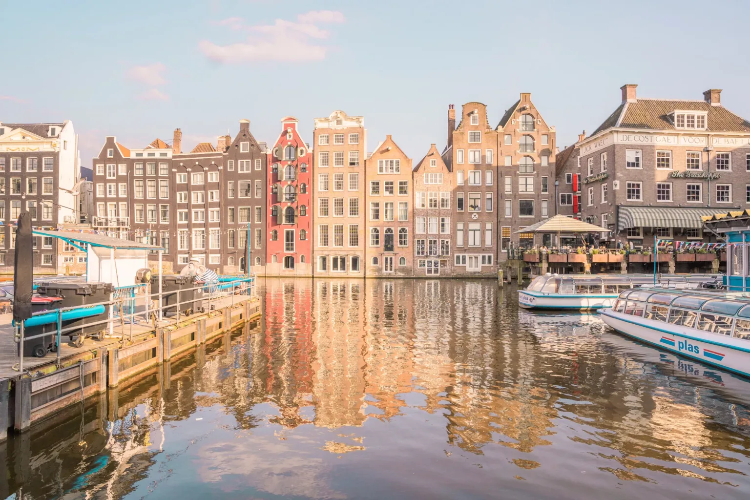 Narrow, gabled canal houses in warm pastel tones line a sunlit Amsterdam waterfront with tour boats moored in the foreground.