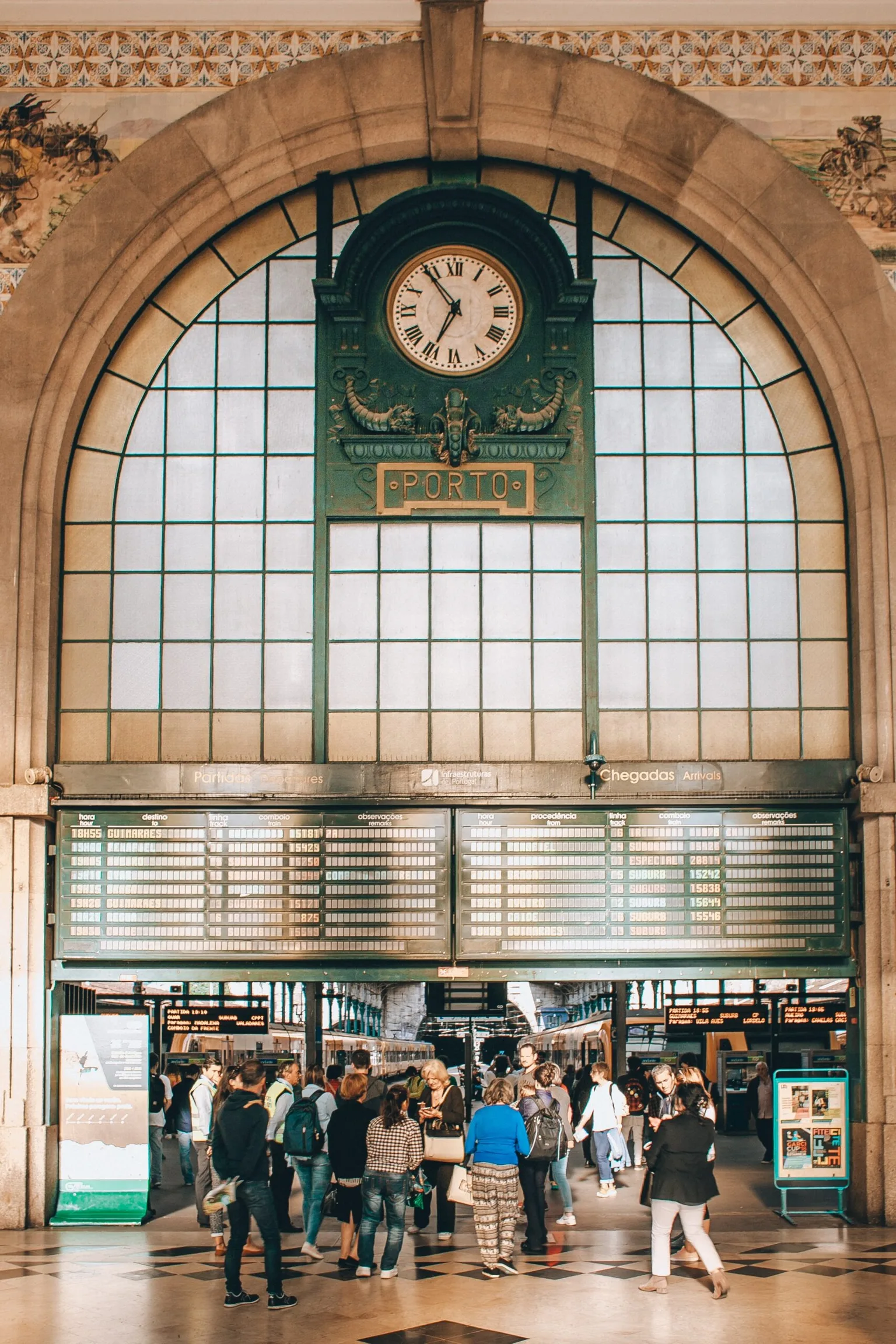 Grand arched station hall with a large clock and departure boards above a crowd of travellers beneath "Porto" sign