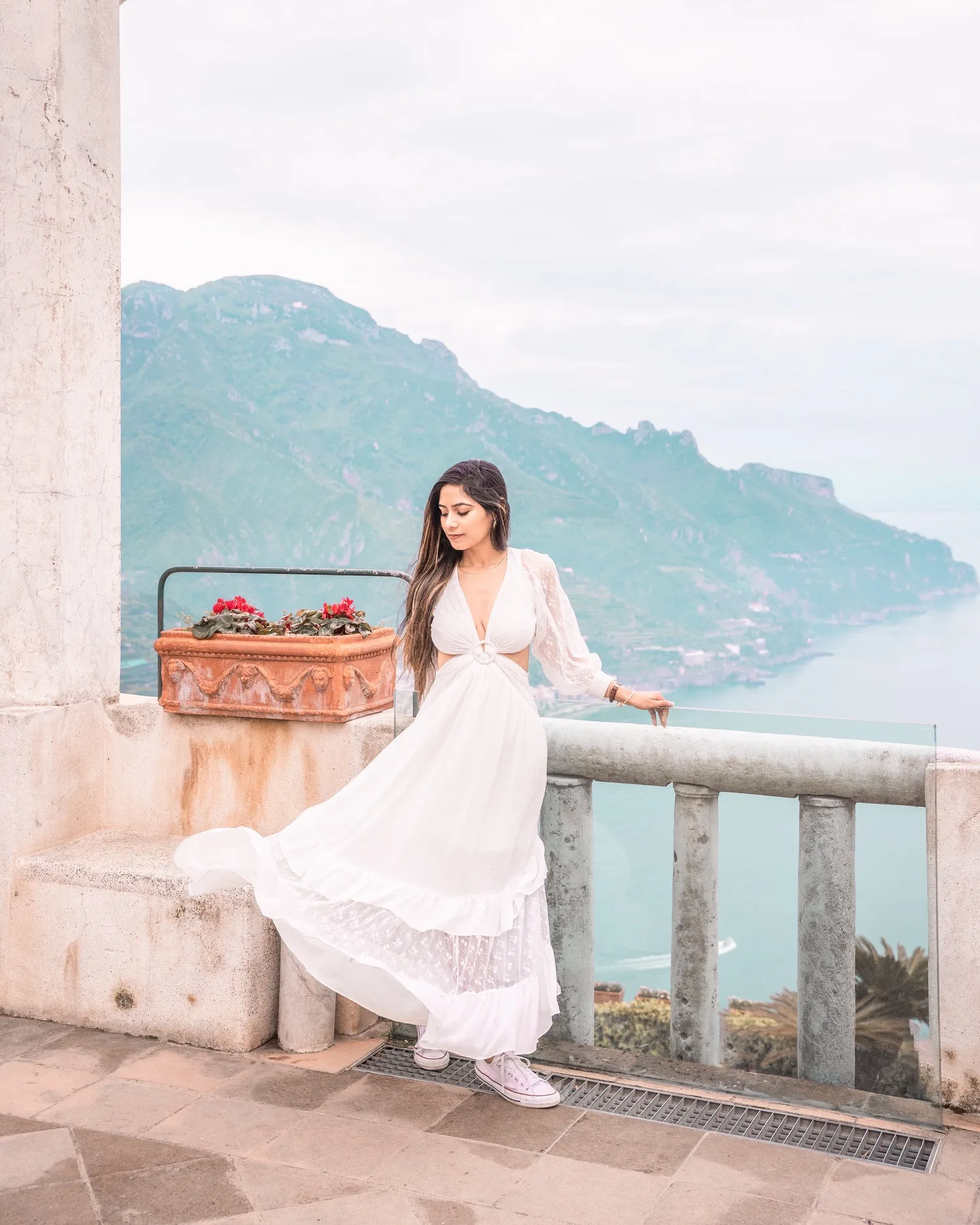 Woman in a flowing white dress and sneakers posing on a stone terrace overlooking coastal cliffs and the sea.