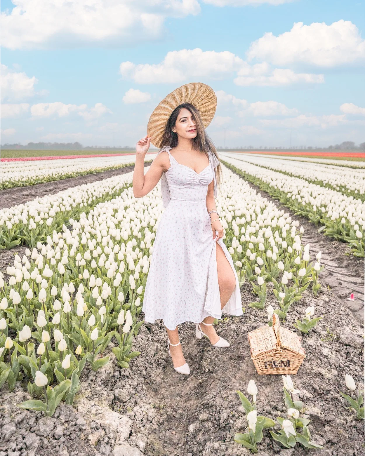 Woman in a white sundress and wide straw hat standing in rows of white tulips with a picnic basket at her feet under a blue sky