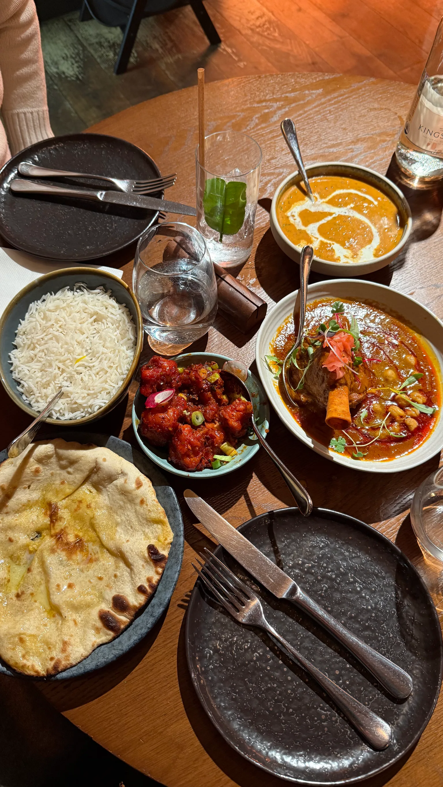 Table set with Indian dishes including naan, basmati rice, spicy cauliflower, butter curry and a lamb shank curry on a wooden table.