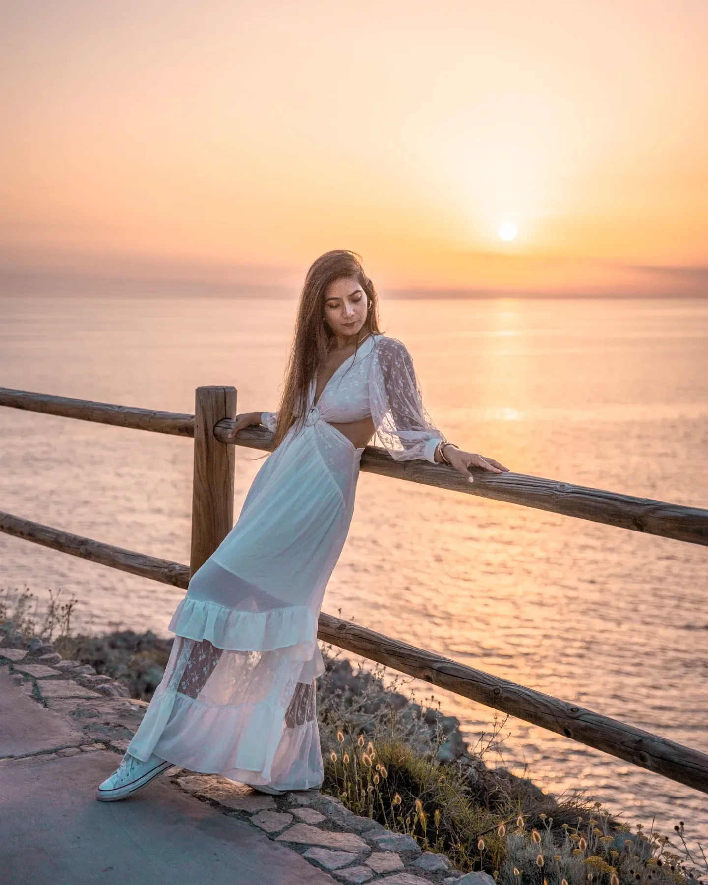 Woman in a flowing white dress leaning on a wooden railing and gazing at a golden sunset over the sea