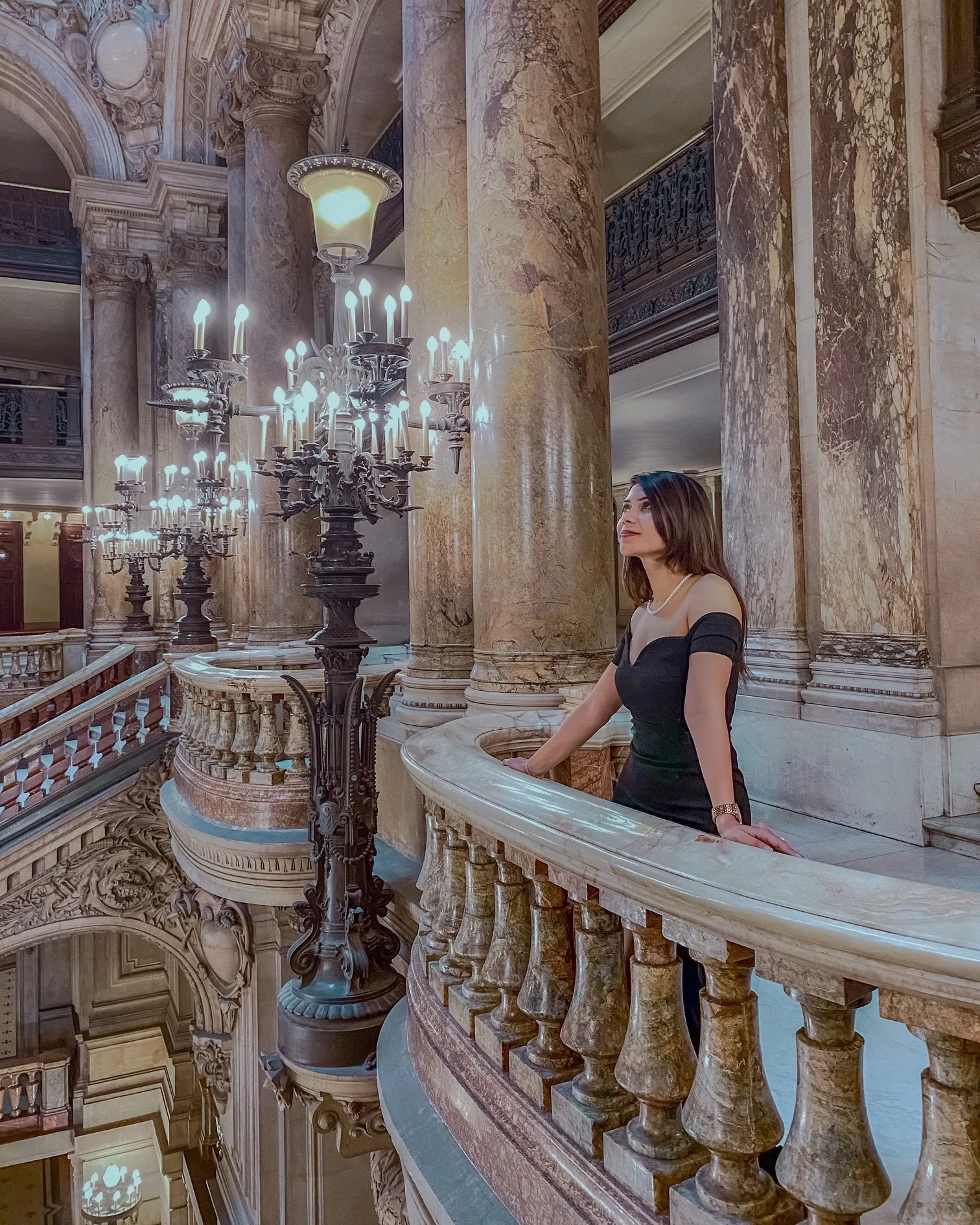 Woman in an elegant black dress gazing across an ornate marble balustrade beneath towering columns and glowing candelabras in a grand historic interior