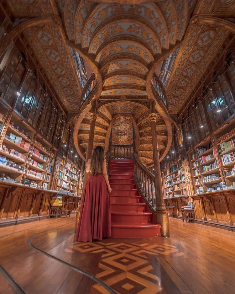 Woman in a flowing red dress ascending a glossy red spiral staircase beneath an ornate carved wooden ceiling inside a grand historic bookstore.
