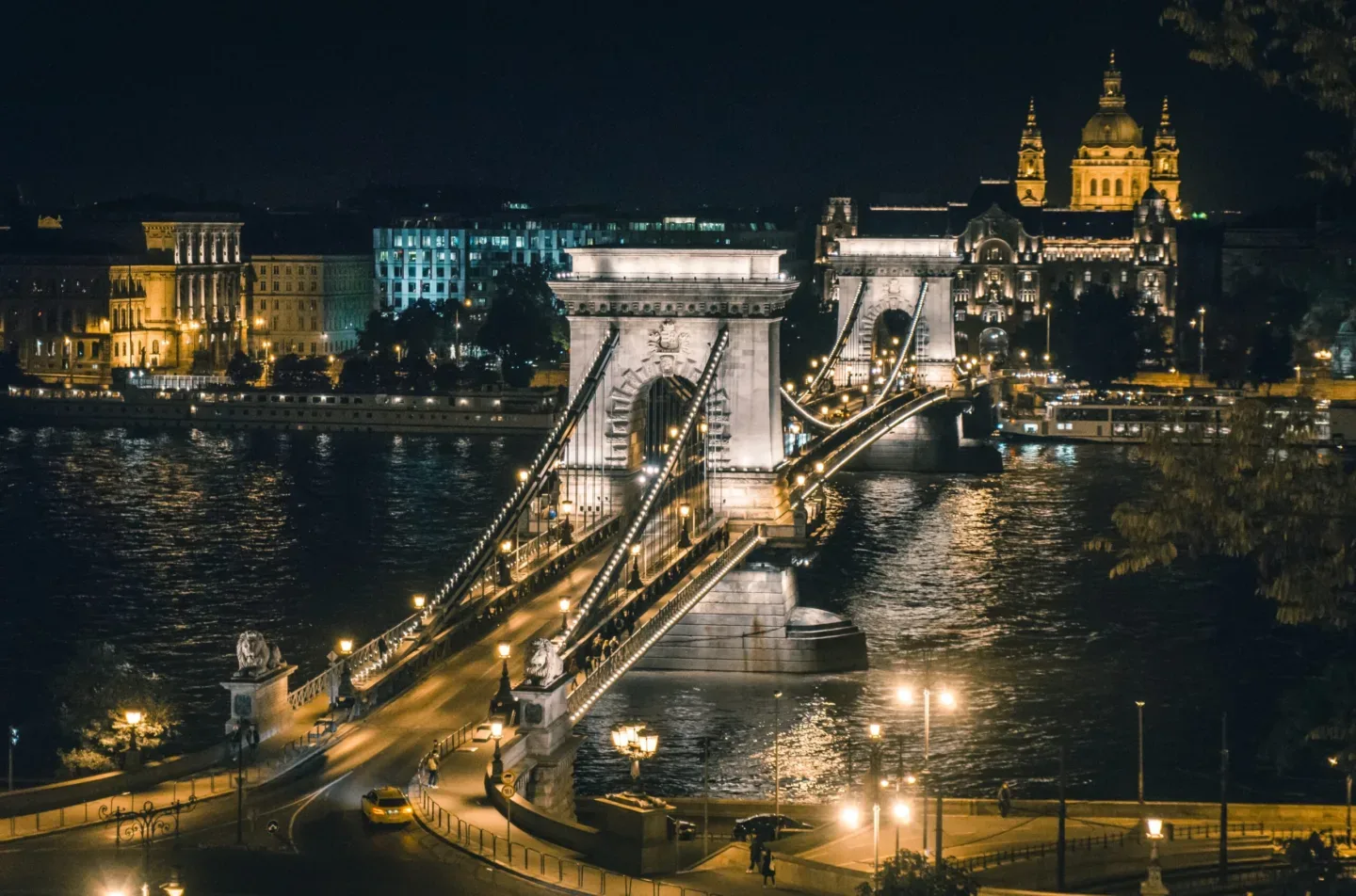 Chain Bridge in Budapest lit by warm streetlights at night, its stone towers and suspension cables reflecting on the Danube.