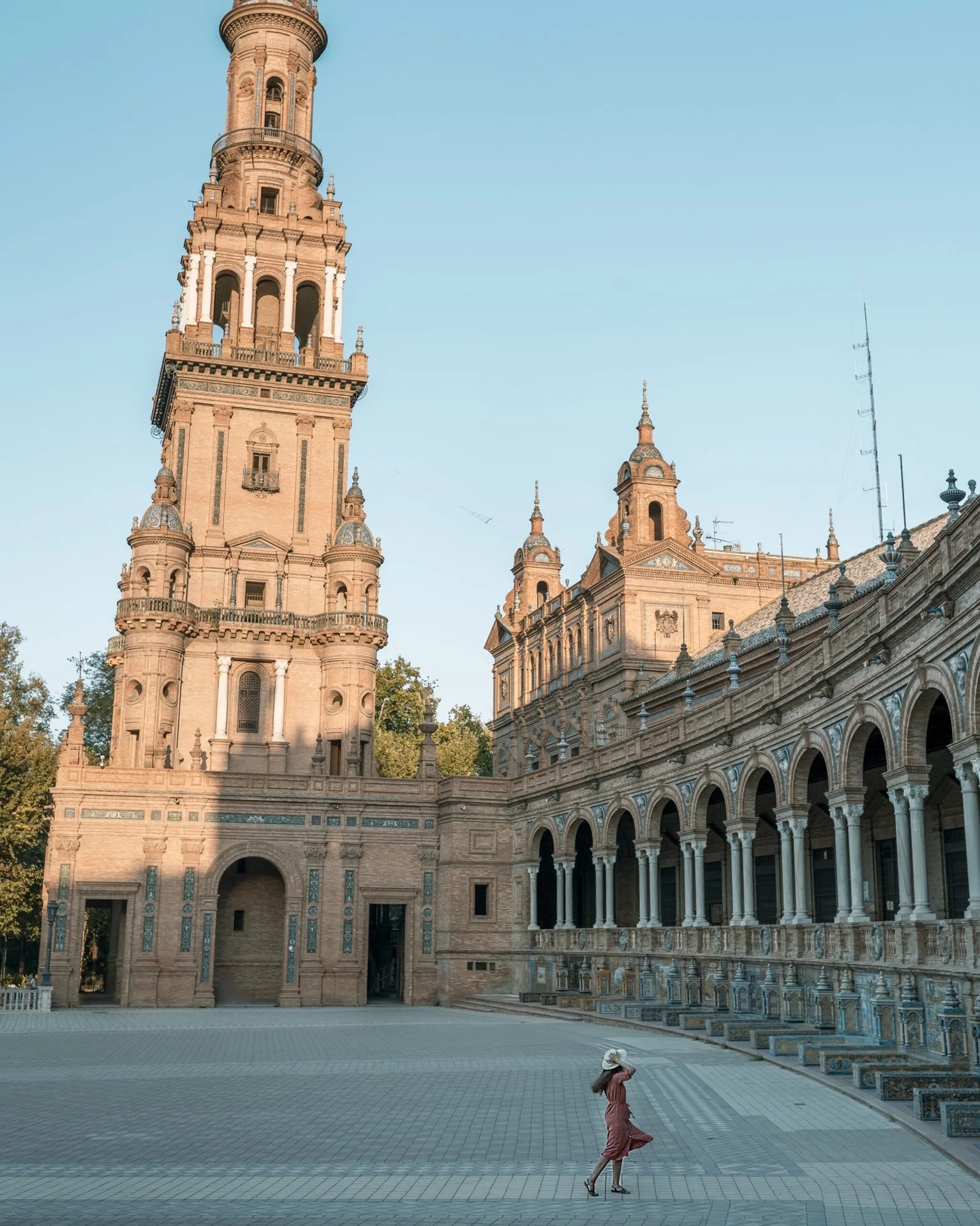 Woman in a pink dress and sunhat walking across the plaza beside the ornate towers and arched colonnade of Plaza de España.