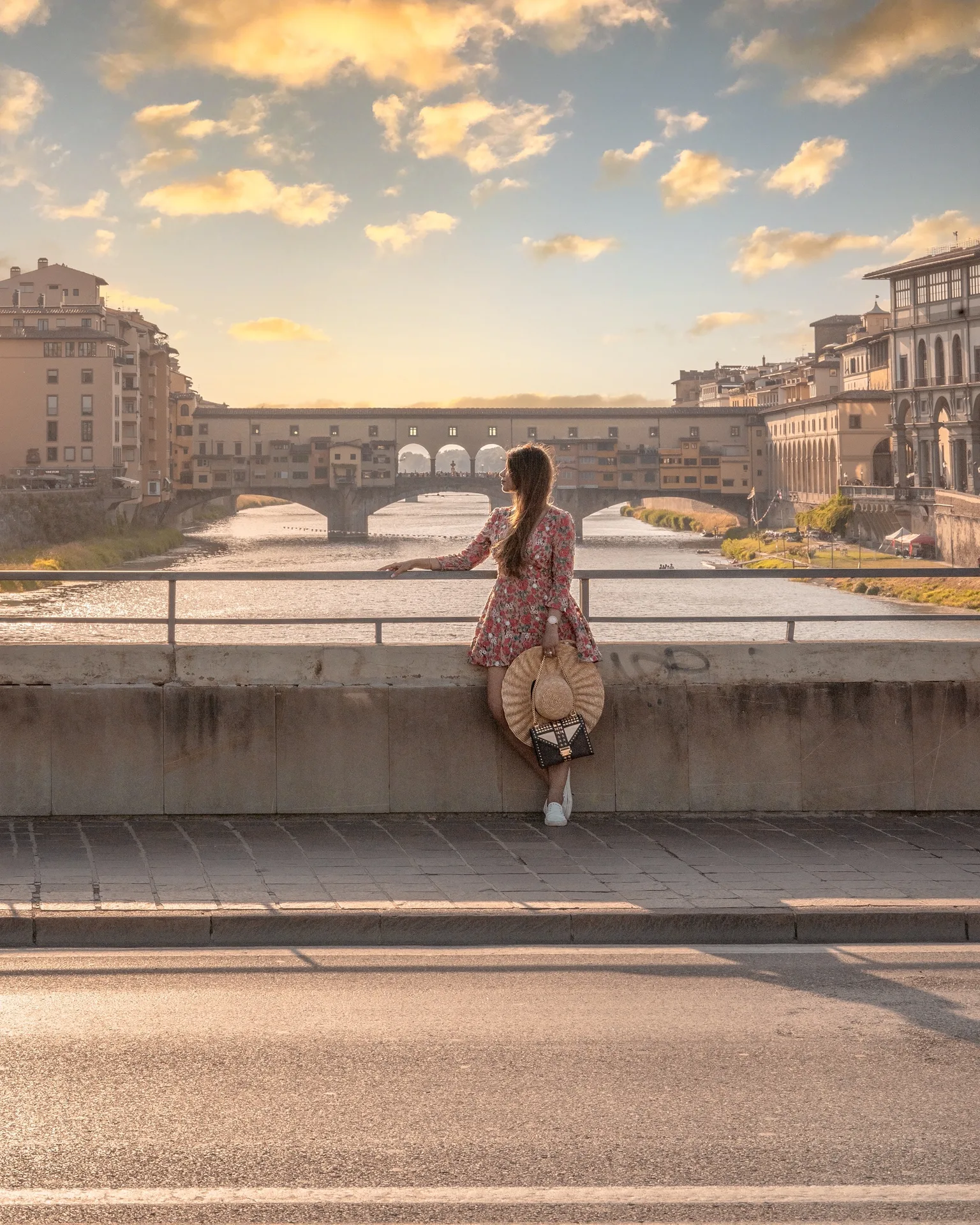 Woman in a floral dress leaning on a bridge railing, looking toward the sunlit Ponte Vecchio over the Arno River at golden hour.
