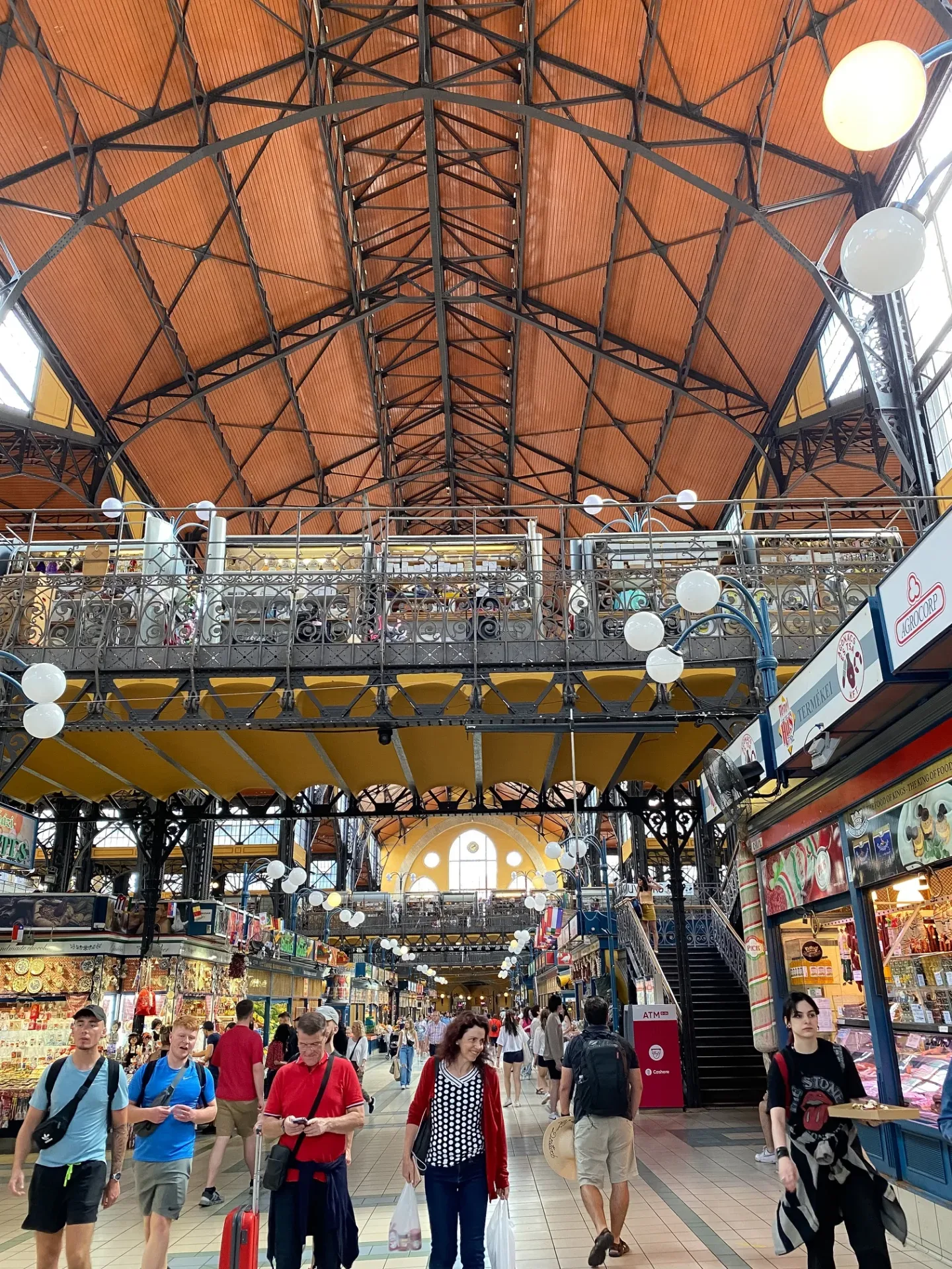 Spacious indoor market hall with ornate iron balconies, high wooden vaulted ceiling and shoppers browsing stalls below.