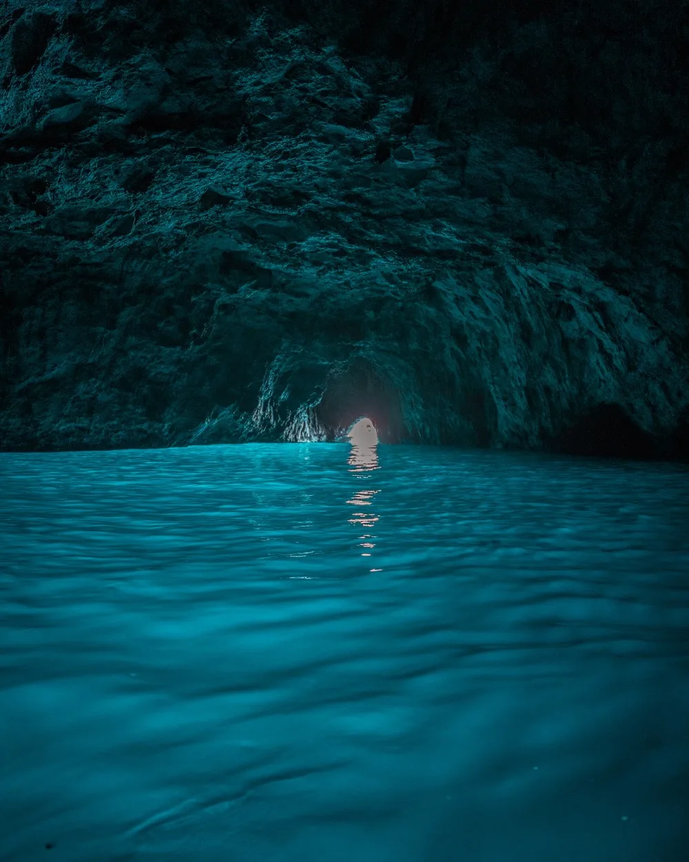 Turquoise seawater inside a dim sea cave with a small bright opening and light reflecting on the rippled surface.