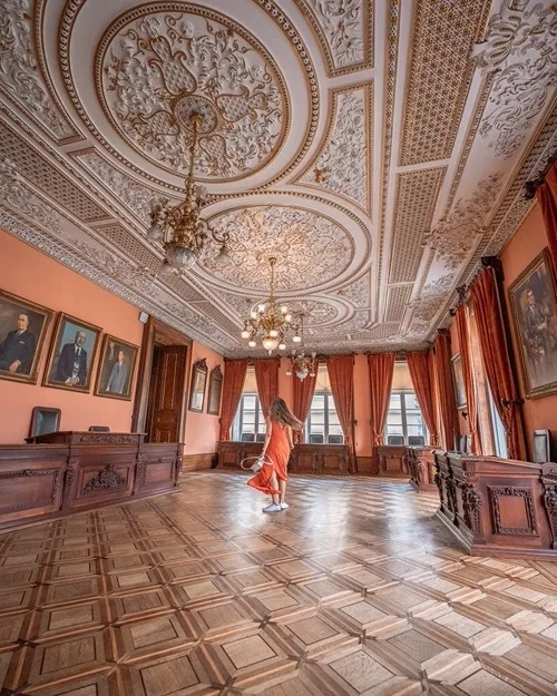 Woman in an orange dress twirling beneath ornate plaster ceilings in a grand, wood-paneled historic reception room.