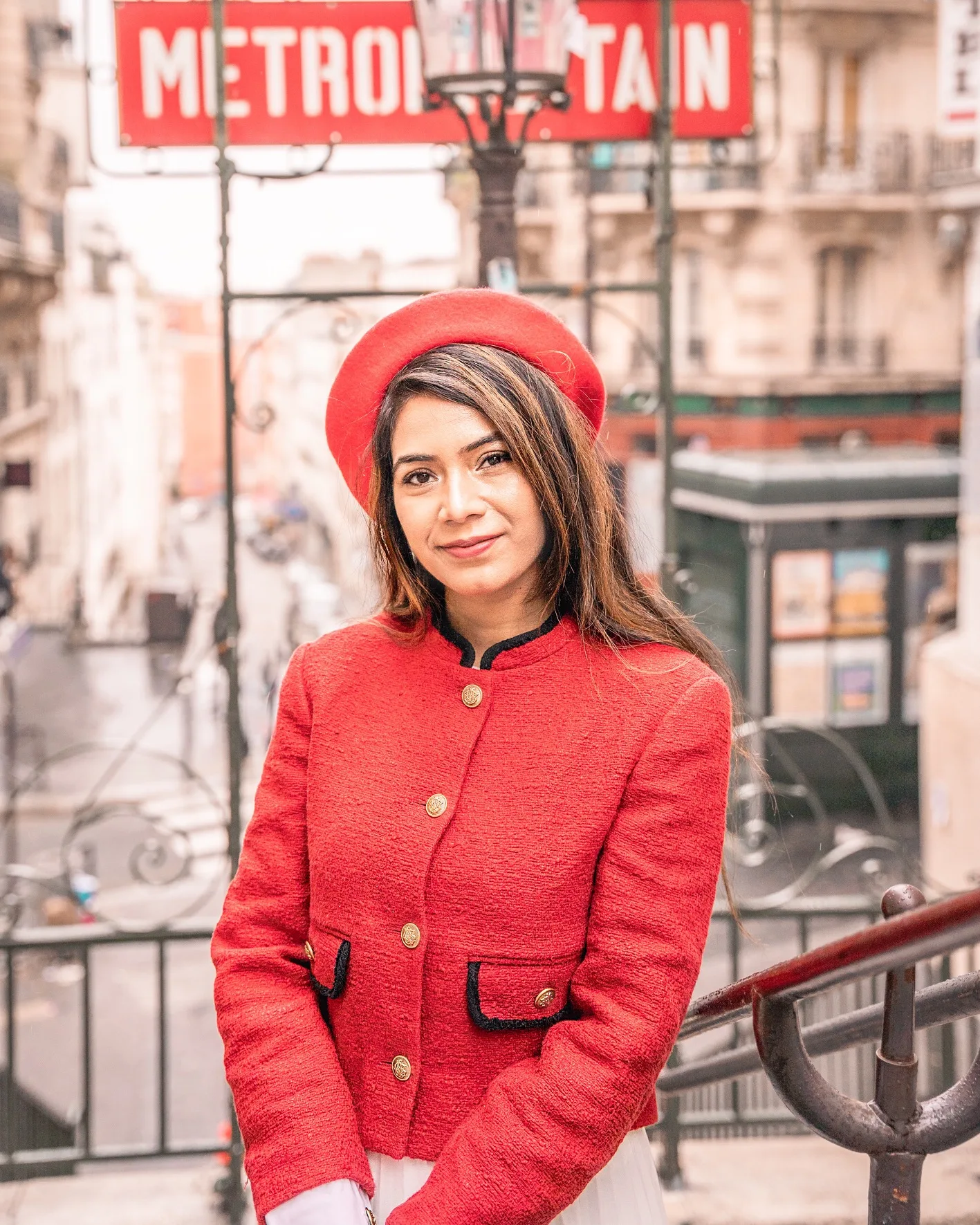 Woman in a red beret and matching coat smiling on the steps of a Paris Métro entrance with the "Metropolitan" sign in the background.