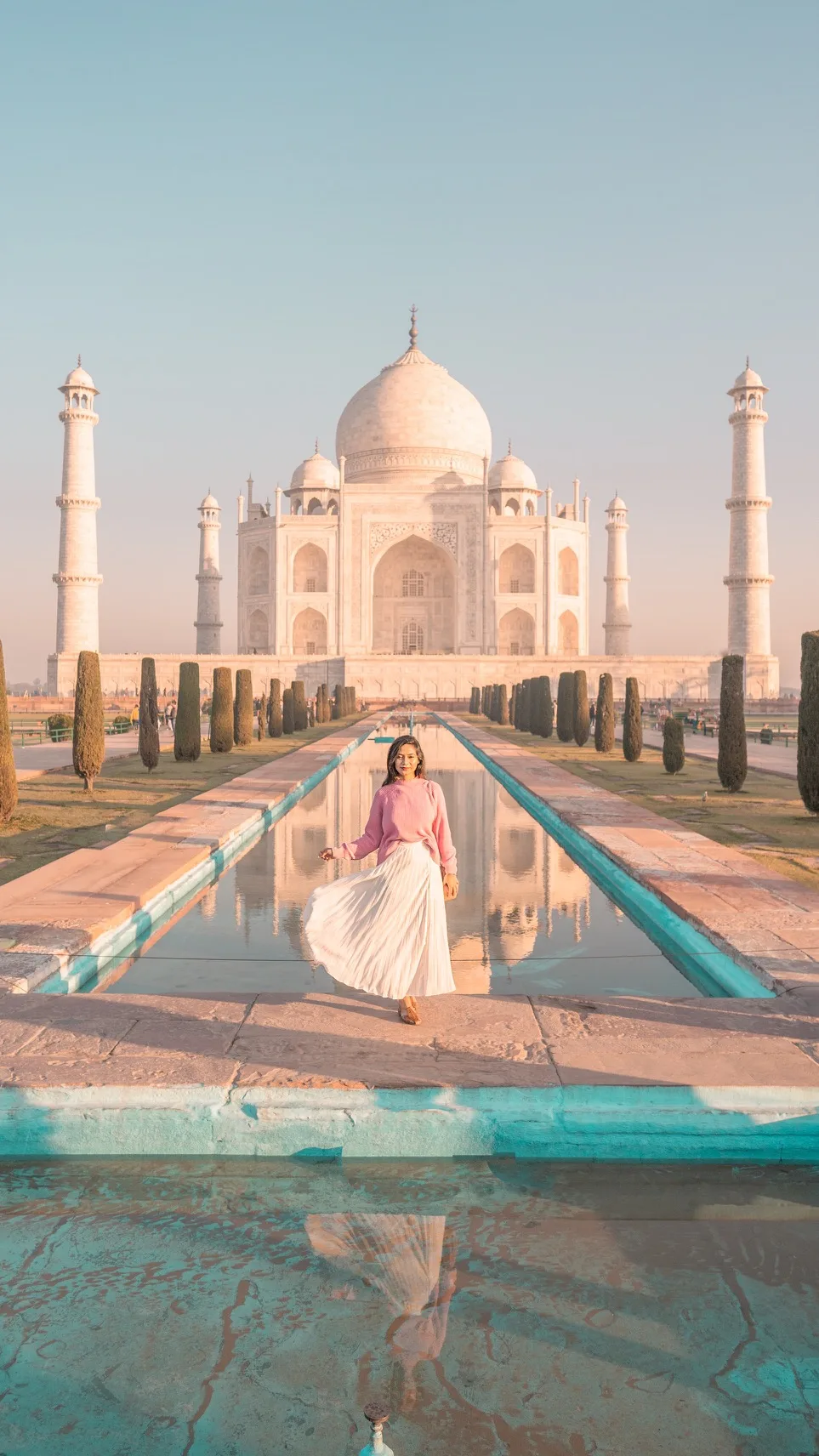 Woman in a pink sweater and flowing white skirt walking beside the reflecting pool with the Taj Mahal in soft morning light