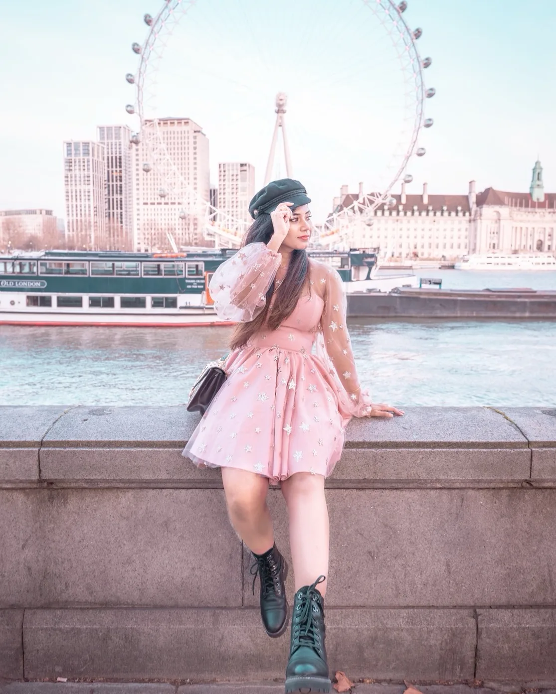 Woman in a pink star-patterned dress and black boots sitting on a riverside wall with the London Eye and riverboats behind her